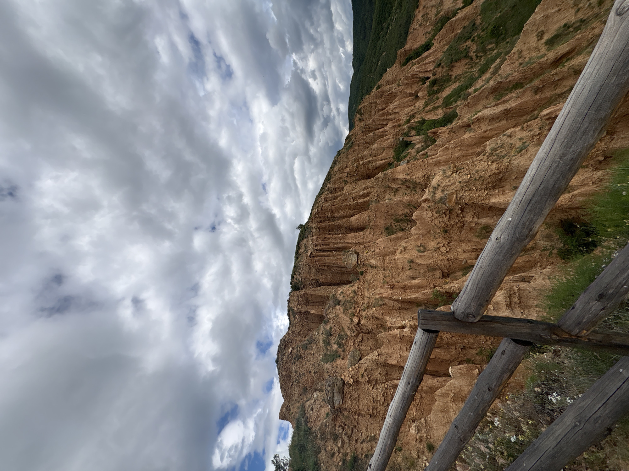 The image depicts the Stob Pyramids in Stob, Bulgaria, viewed from behind a wooden fence. The rock formations are a reddish-brown color with vertical striations. Sparse green vegetation grows on the pyramids and adjacent to the fence. The sky is filled with numerous gray clouds.
