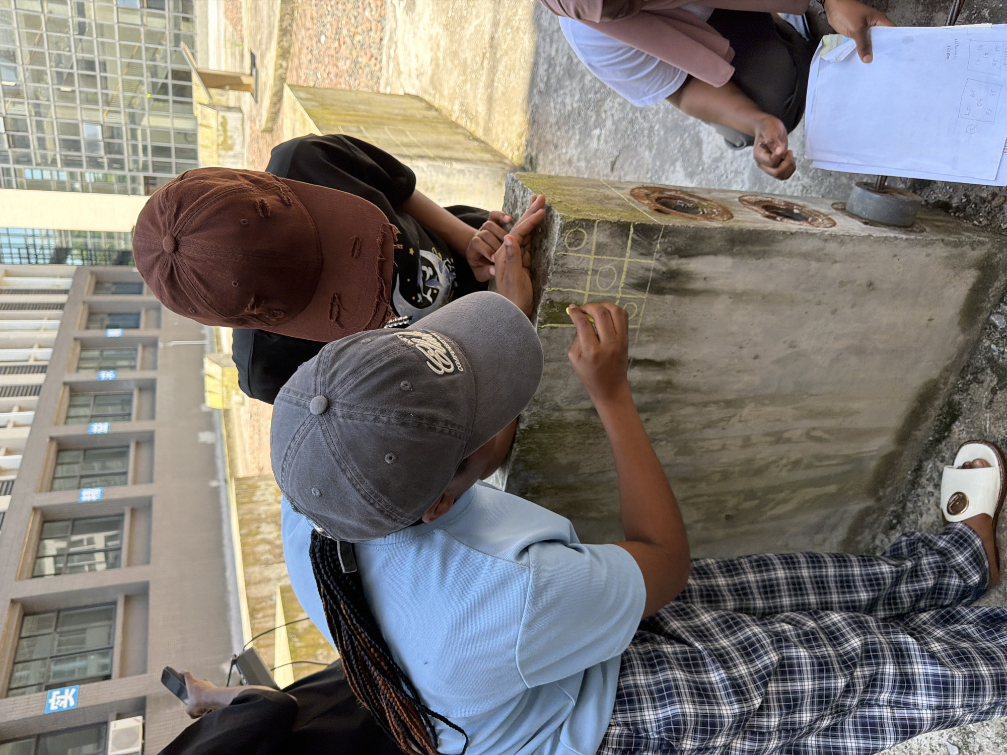 In the city of Jiangjin, China, three individuals are depicted outdoors engaged in an activity. Two of them, wearing baseball caps, are leaning over a concrete structure. One, in a gray cap and blue shirt, is drawing with chalk on the structure, which has a grid of squares and circles drawn on it. The other, in a brown cap and black shirt, is pointing at the drawn grid. A third individual, partially visible, is kneeling to their right, holding papers. The environment suggests an urban setting with a modern building in the background. The concrete structure appears to be part of some sort of outdoor fixture.