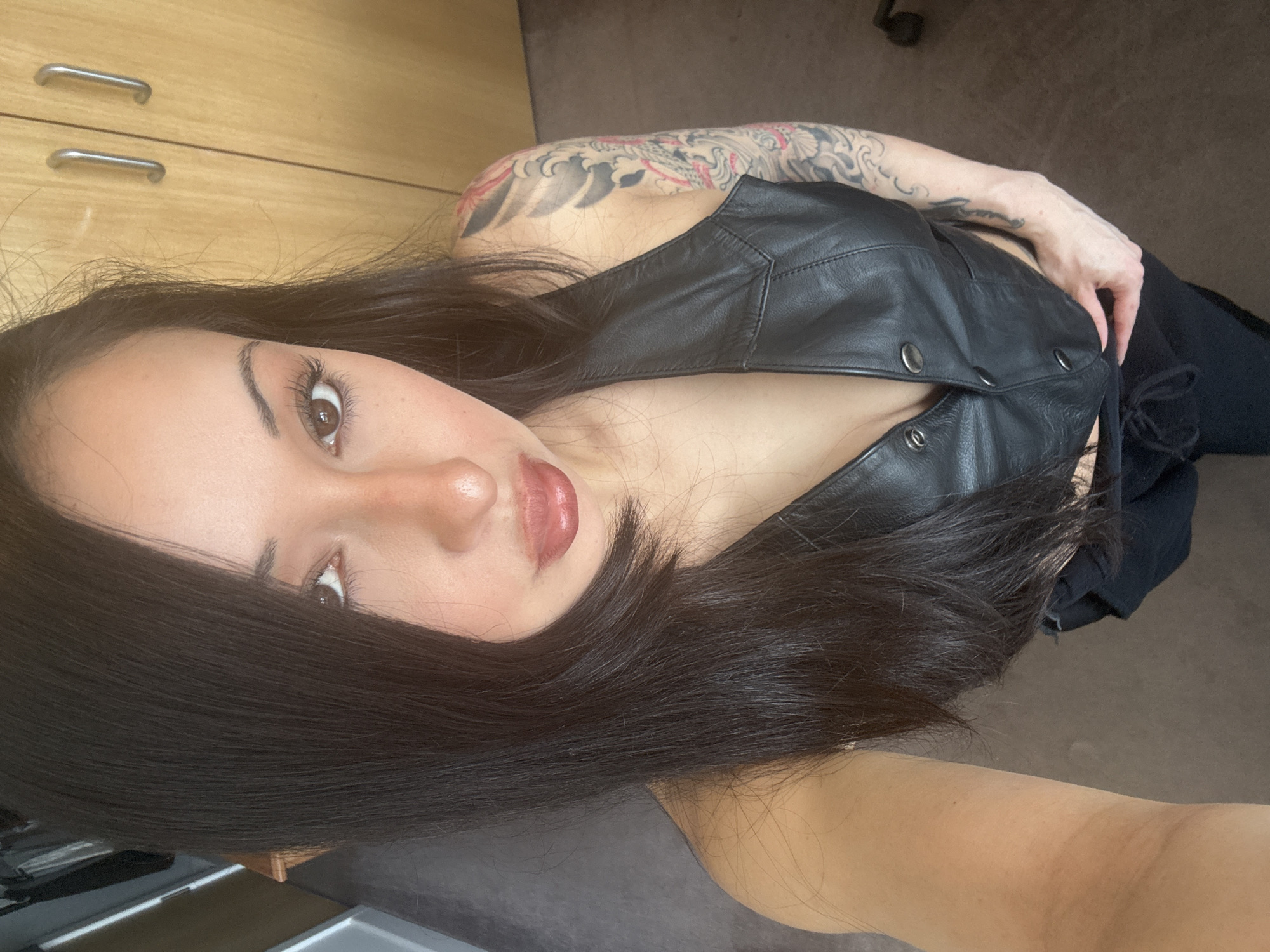 A young woman is the central subject of this close-up selfie, captured from a high angle looking down at her. She has long, dark, straight hair that falls around her face and shoulders. Her facial features include large brown eyes, prominent eyebrows, and lips adorned with a shiny, light red or nude-toned lip gloss. She has visible eyeliner and mascara.
She is wearing a black, leather-look vest or top with silver snap buttons down the front. A large, intricate tattoo sleeve featuring what appears to be traditional Japanese-style waves and possibly a dragon or similar mythical creature is visible on her right arm. Her left arm and hand are also visible, resting near her side, and she appears to be wearing dark pants or leggings.
The background suggests an indoor setting, likely a room with wooden cabinets or drawers featuring silver metal bar handles. A dark floor or carpet is also visible. The lighting is bright and natural, indicating it is likely daytime. The overall mood conveyed is calm and confident. There is no visible text in the image. The location is in London, United Kingdom.