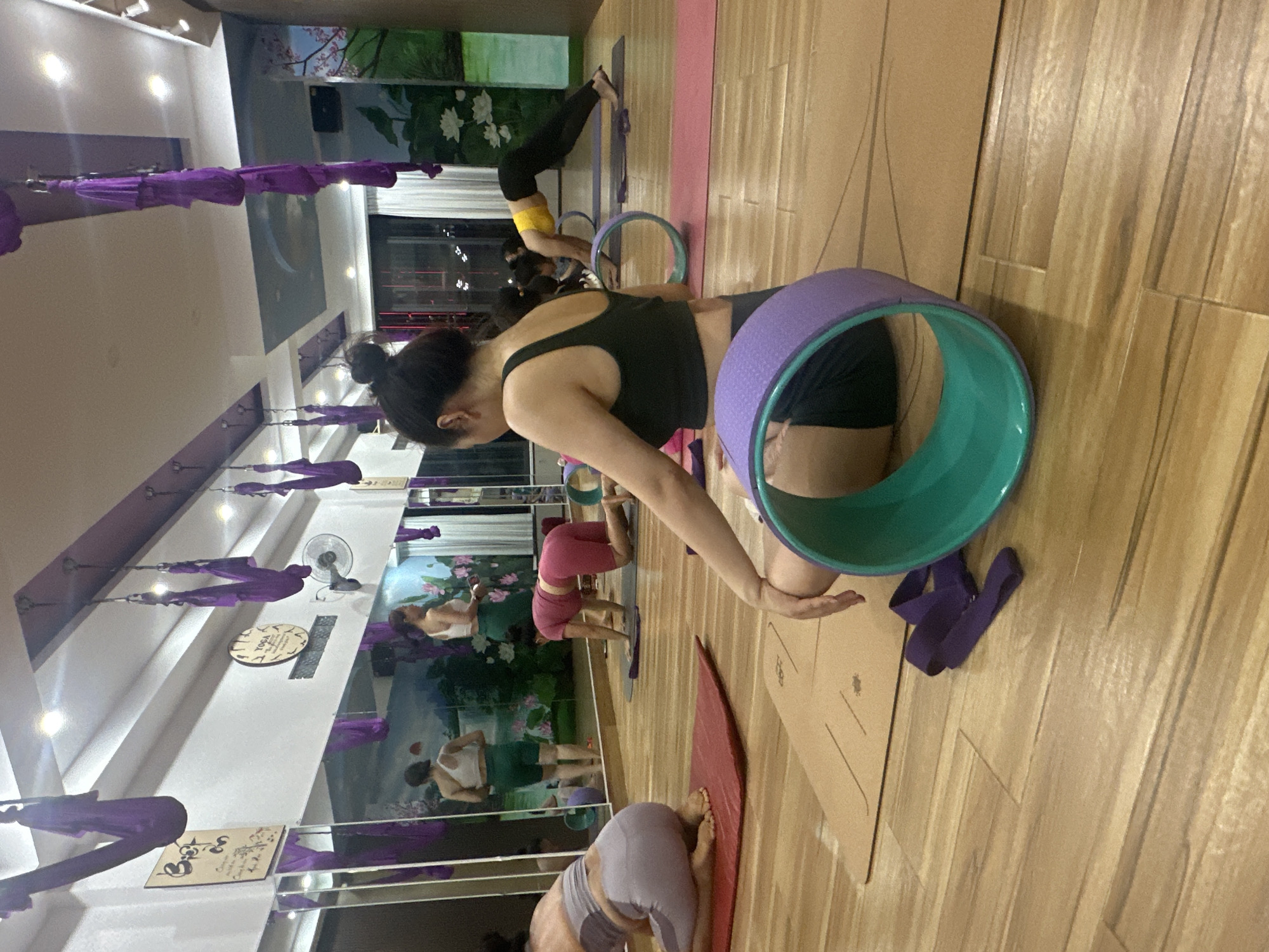 A group of women is engaged in a yoga class within a studio. The studio features wooden flooring, mirrors along one wall, and a mural depicting a landscape with flowers and trees. Purple aerial yoga hammocks are suspended from the ceiling. In the foreground, a woman is seated on a yoga mat, facing away from the camera, with a large purple and teal yoga wheel beside her. Another woman in a pink outfit is on her hands and knees on a yoga mat in the middle ground. Further back, a woman in a yellow top and black pants is in a downward-facing dog pose with a yoga wheel. Two other women are visible in the reflections of the mirrors, one wearing a white top and green shorts, and another in a similar outfit. The class appears to be taking place in the city of Bac Nha Trang Ward, Vietnam.