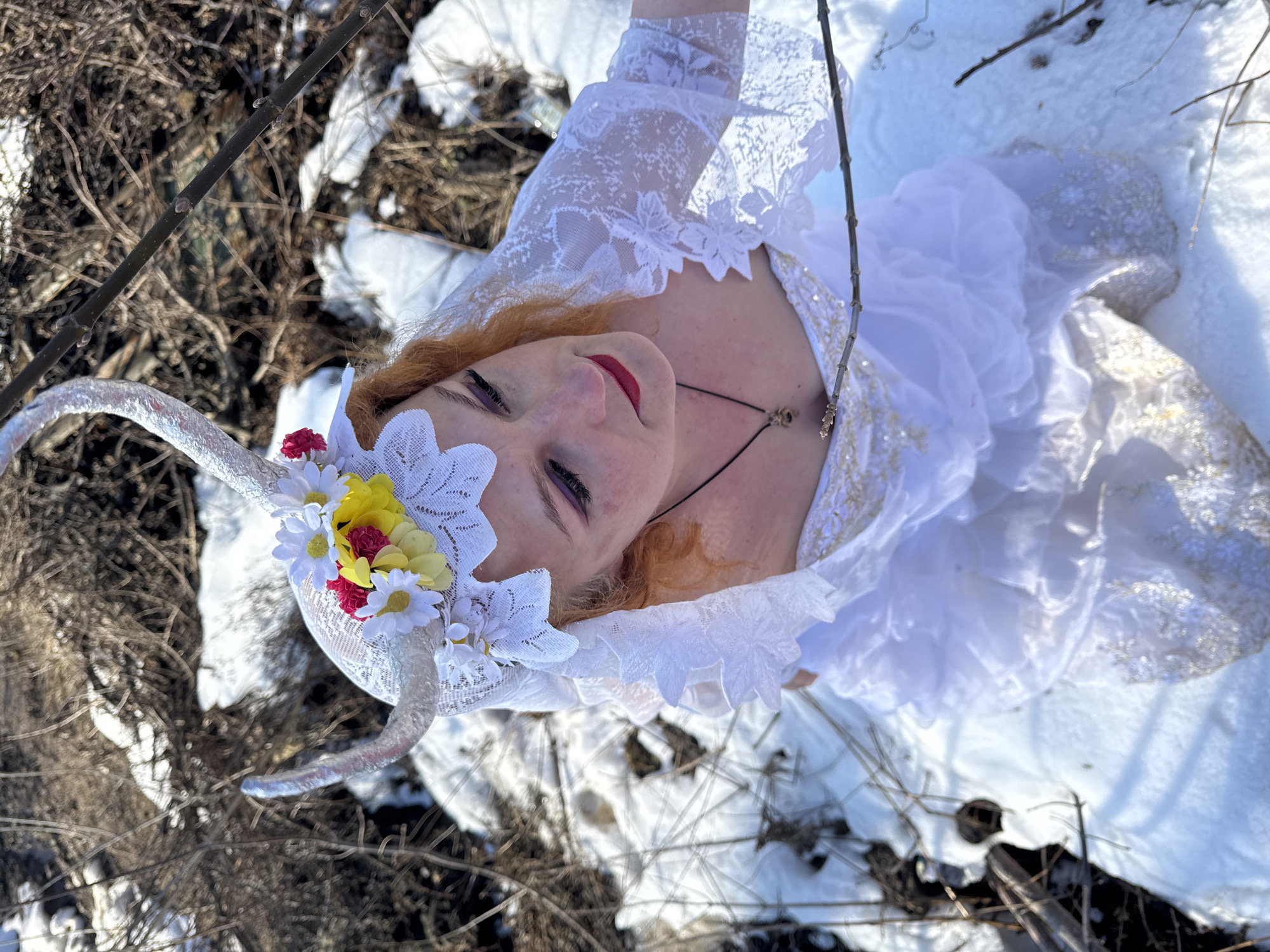A person wearing a white dress and a horned headdress is lying on snow-covered ground amidst dry vegetation. The person has red hair and is wearing red lipstick. The location appears to be outdoors in a natural setting, potentially near Chaikovsky, Russia, given the reference. Specific actions and interactions are not discernable beyond the person lying on the ground. The focus is on the person and their attire in relation to the wintery environment.
