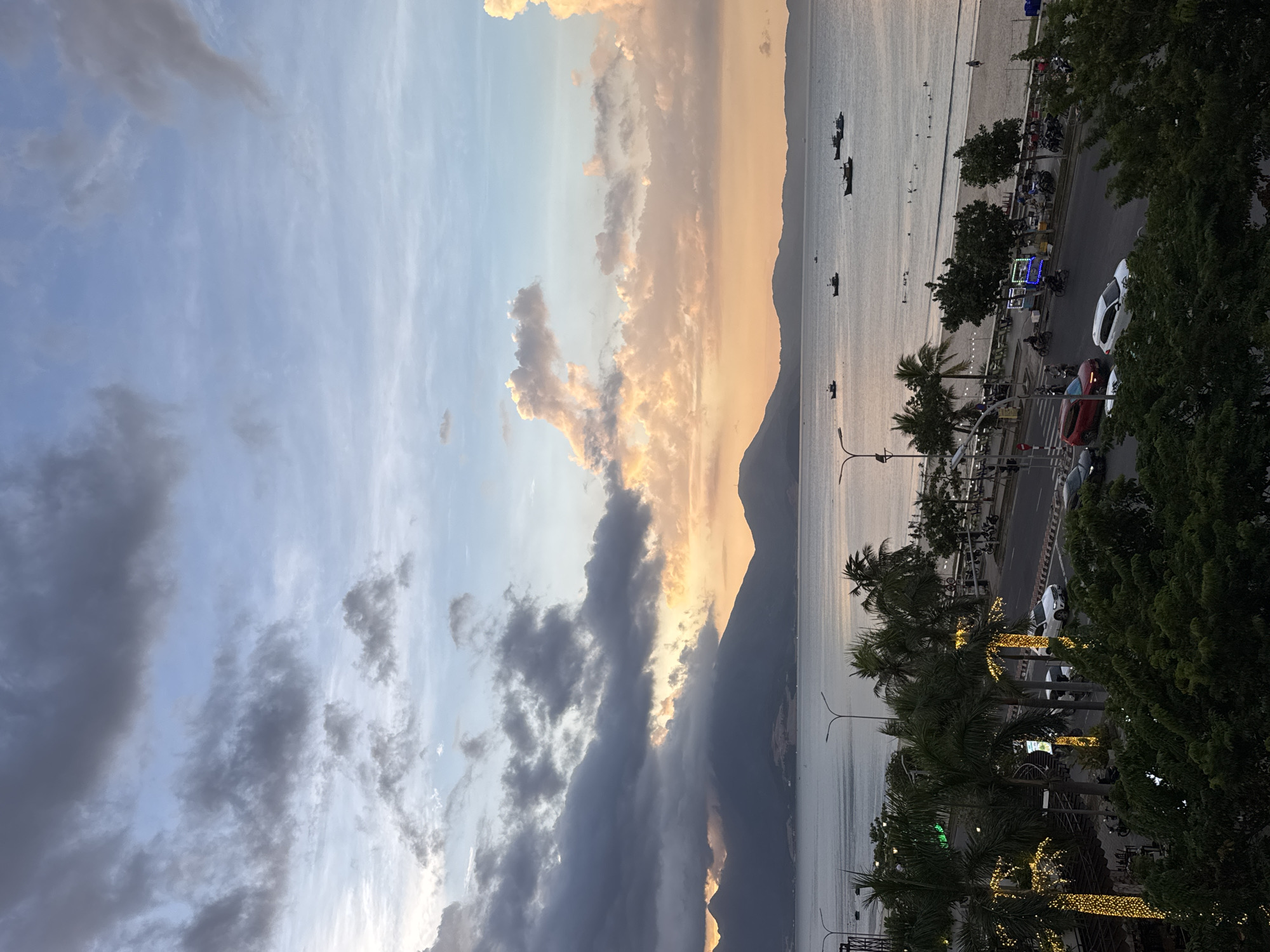 This is an elevated, wide-angle photograph of a coastal city scene at dusk, likely in Đà Nẵng, Vietnam. The sky is filled with a mix of dark, dramatic clouds and lighter, pastel-colored clouds illuminated by the setting sun. The sea is calm, with several small boats scattered across its surface. In the distance, a range of dark mountains forms a silhouette against the horizon.
The middle ground of the image features a beach with a few people visible, bordering a street lined with palm trees. Some of these palm trees are decorated with strings of yellow lights. The street itself is busy with traffic, including cars, motorcycles, and scooters. There are also streetlights and some roadside stalls or businesses.
In the foreground, lush green trees obscure the bottom portion of the frame, suggesting the photograph was taken from a building or a high vantage point. The overall atmosphere is one of a coastal city transitioning from day to night, with a blend of natural beauty and urban activity.