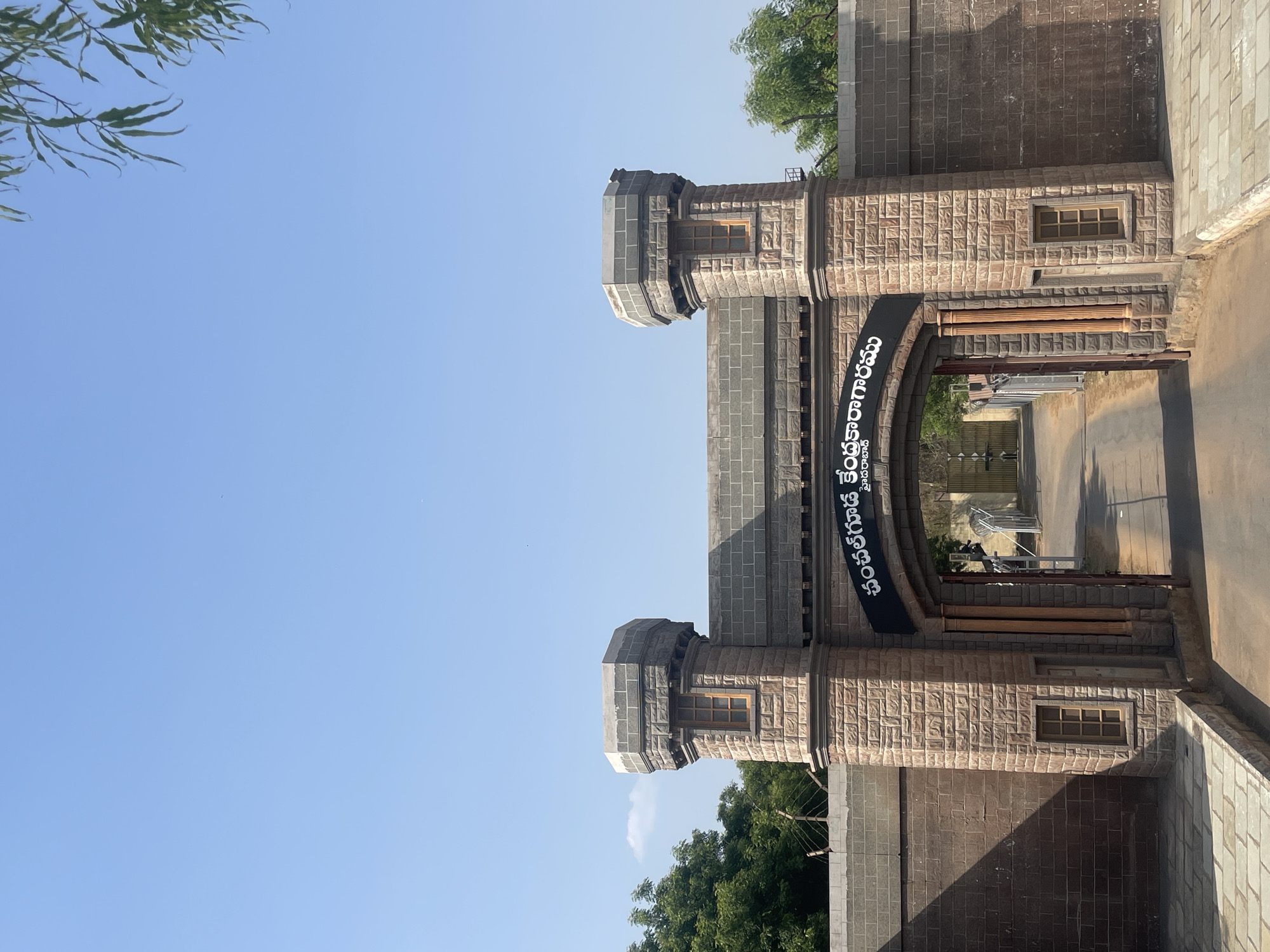The image shows the entrance to the Central Jail in Anajpur, India. The entrance is a stone archway with two towers on either side. A sign in a Telugu script is located above the archway. The view through the archway reveals a path leading into the jail complex. The sky is blue, and there are trees visible in the background. The scene appears to be taken during daylight hours.