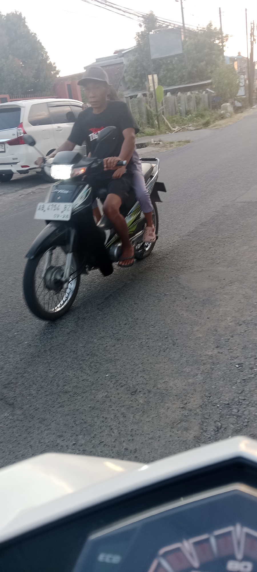 In Maguwoharjo, Indonesia, two individuals are riding a black motorcycle on a paved road. The driver, a male wearing a black t-shirt and a dark cap, is seated in front. A younger person, likely a child, is seated behind the driver, wearing a grey top and light-colored leggings, with pink footwear. The motorcycle's license plate is visible and reads "AB 4754 BZ". In the background, a white vehicle is parked on the side of the road, and several buildings and trees are present. The lighting suggests it is either early morning or late afternoon.