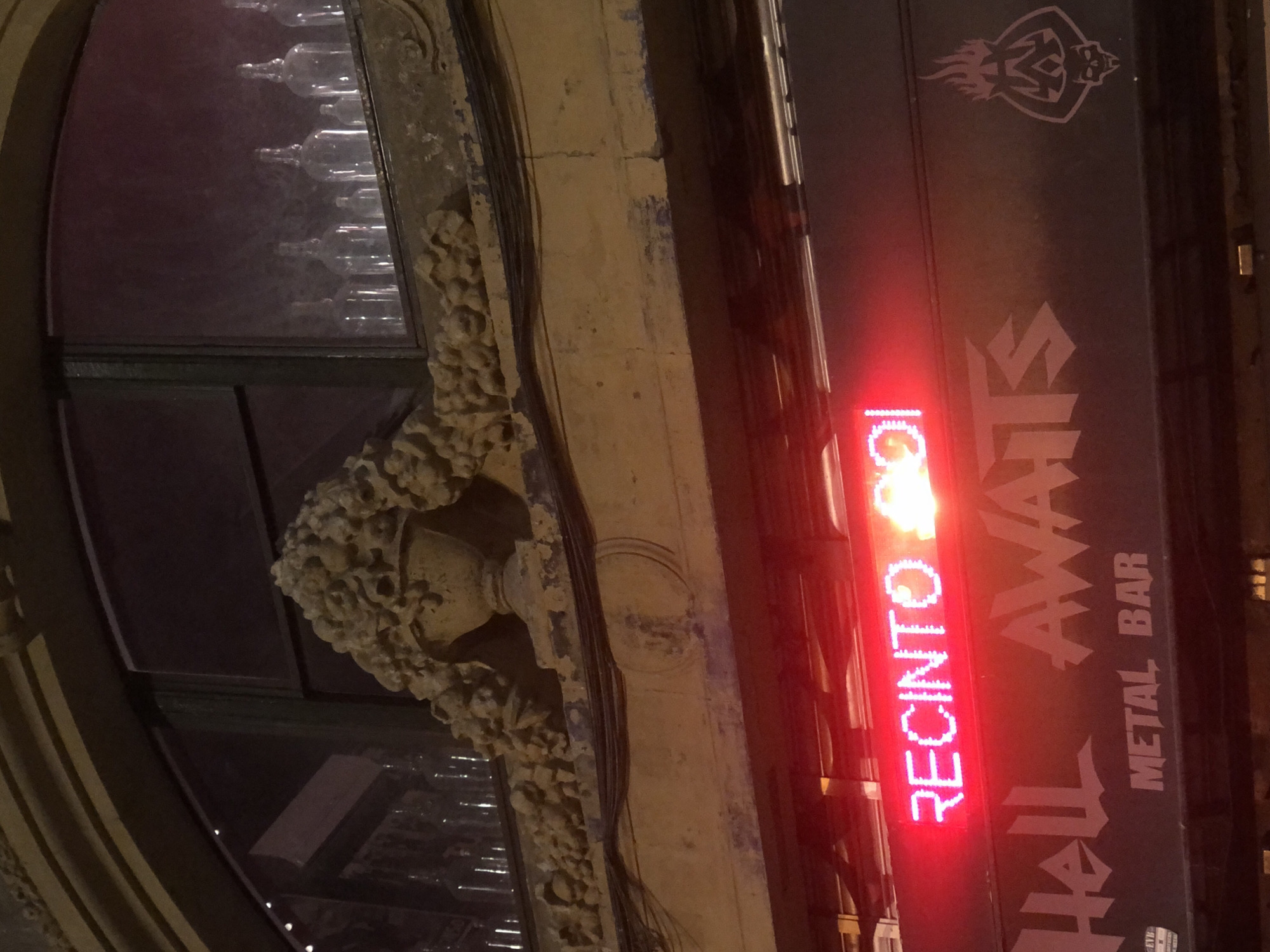 The image shows a nighttime exterior view of a building with a neon sign and decorative architectural elements. The neon sign is red and displays the text "RECINTOS" followed by some unreadable characters. Below the neon sign, a dark sign features stylized white text that reads "Hell AWATS" and "METAL BAR." To the right of the "AWATS" text, there is a graphic of a flaming skull. Above the signs, there is a stucco facade with ornate, detailed carvings that appear to depict a vase overflowing with foliage or flowers. Behind the carvings, a window is visible, through which several clear glass bottles can be seen lined up. The building's architecture suggests an older structure. The environment is dimly lit, as expected for nighttime. No people are visible in the image. The location is unknown.