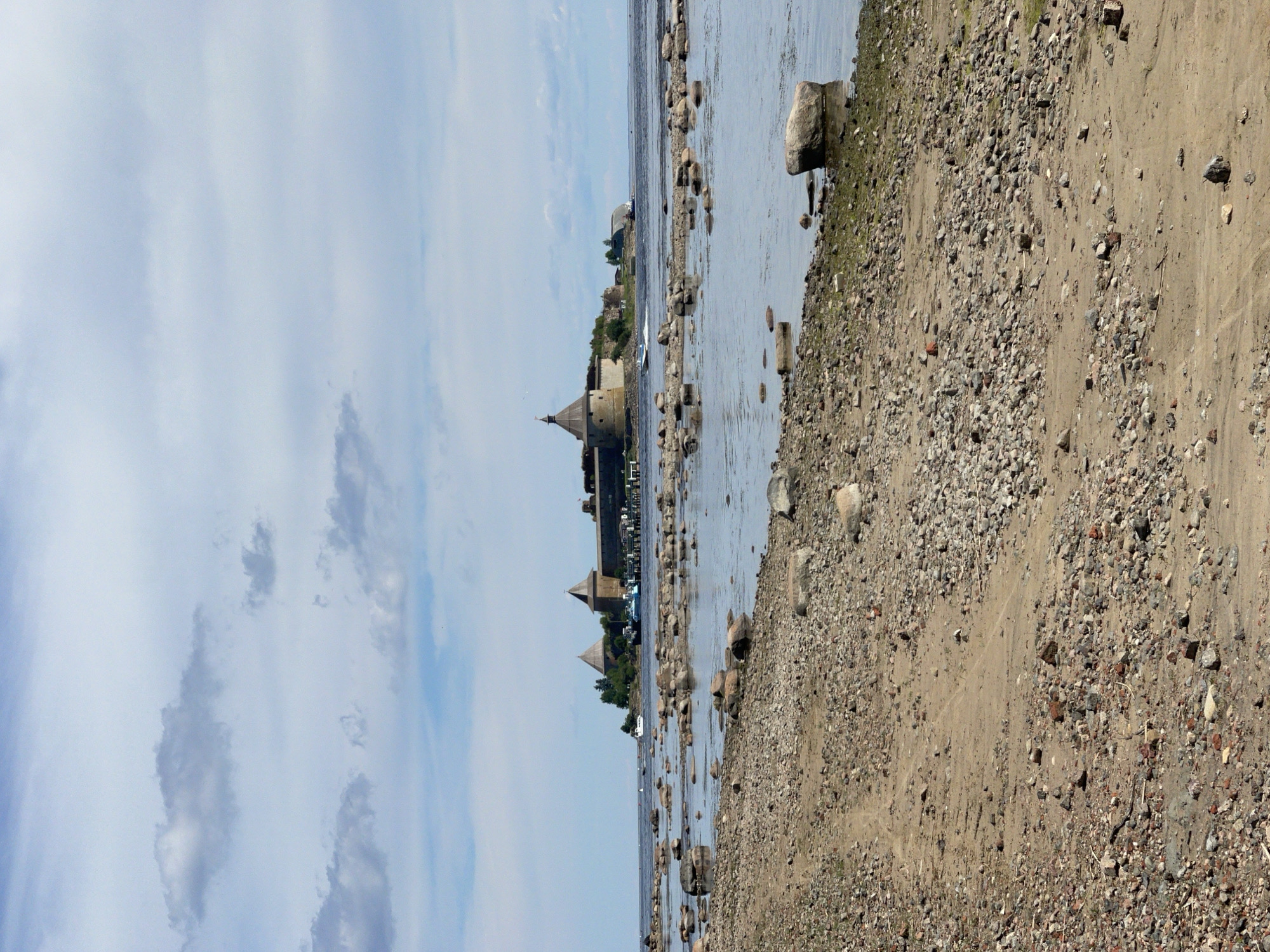 The image depicts the Oreshek Fortress, a historical fortress located on the island of Oreshek in Shlisselburg, Russia. The fortress, with its distinctive conical-roofed towers and stone walls, is situated on a small, tree-covered island in the middle of a body of water, likely Lake Ladoga. The foreground of the image shows a rocky, sandy shoreline with scattered stones and vegetation. The water is calm, with a few boats visible in the distance, including a white speedboat creating a wake as it moves across the water. The sky is partly cloudy with a mix of blue sky and white cumulus clouds. The overall scene conveys a sense of historical grandeur and natural beauty.