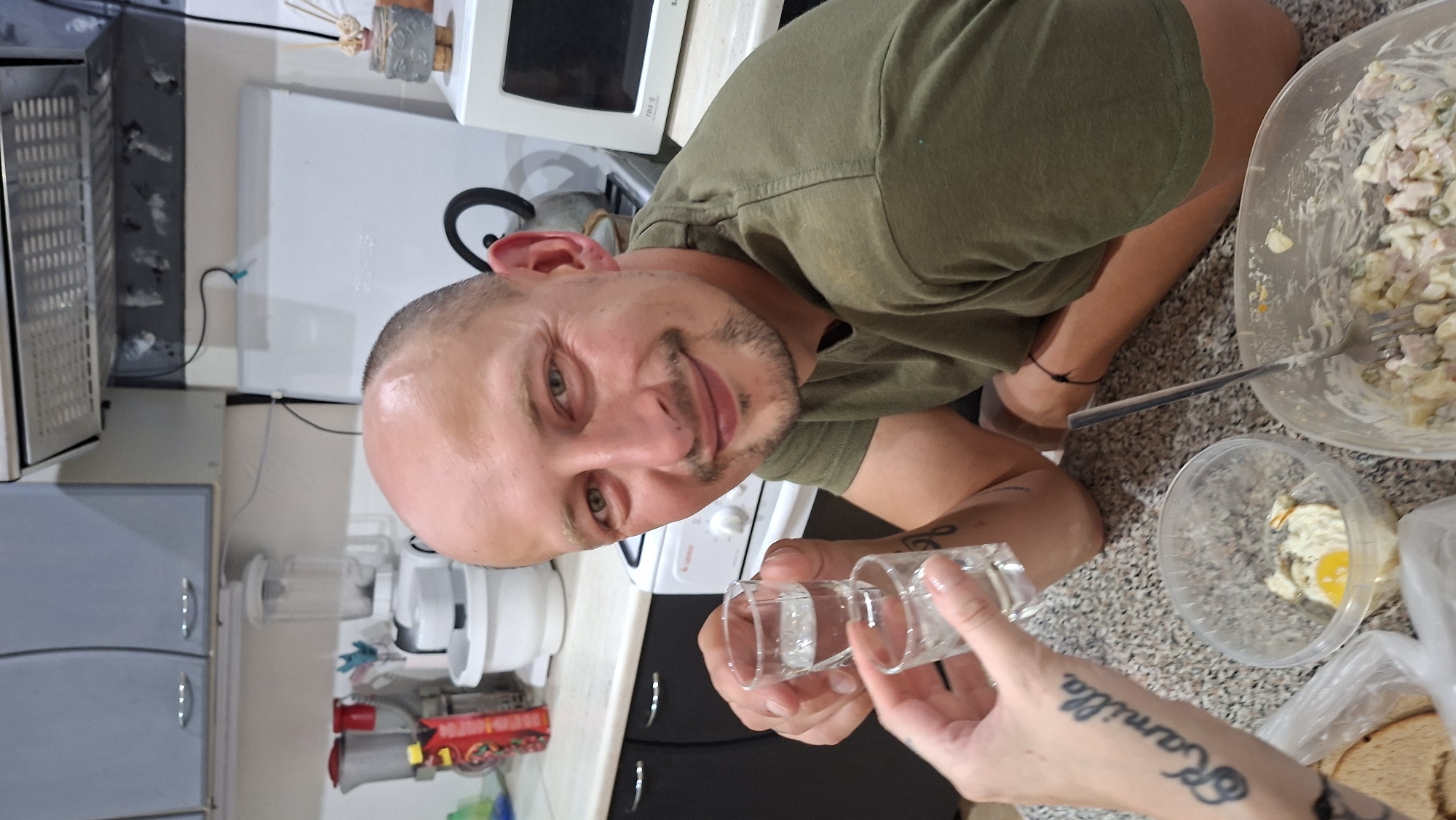 The image depicts a medium shot of a man with a shaved head, a mustache, and a goatee, smiling as he holds up two shot glasses of clear liquid. He is wearing a green t-shirt. His left hand, with a tattoo reading "Kamilla" on the forearm, is also visible holding a shot glass. In the background, a kitchen counter is visible with various objects including a white food processor, a microwave oven, a blender, and containers of food. The man is seated, with his legs crossed. The overall setting appears to be a residential kitchen. The image is taken from a slightly low angle, focusing on the man and the drinks.