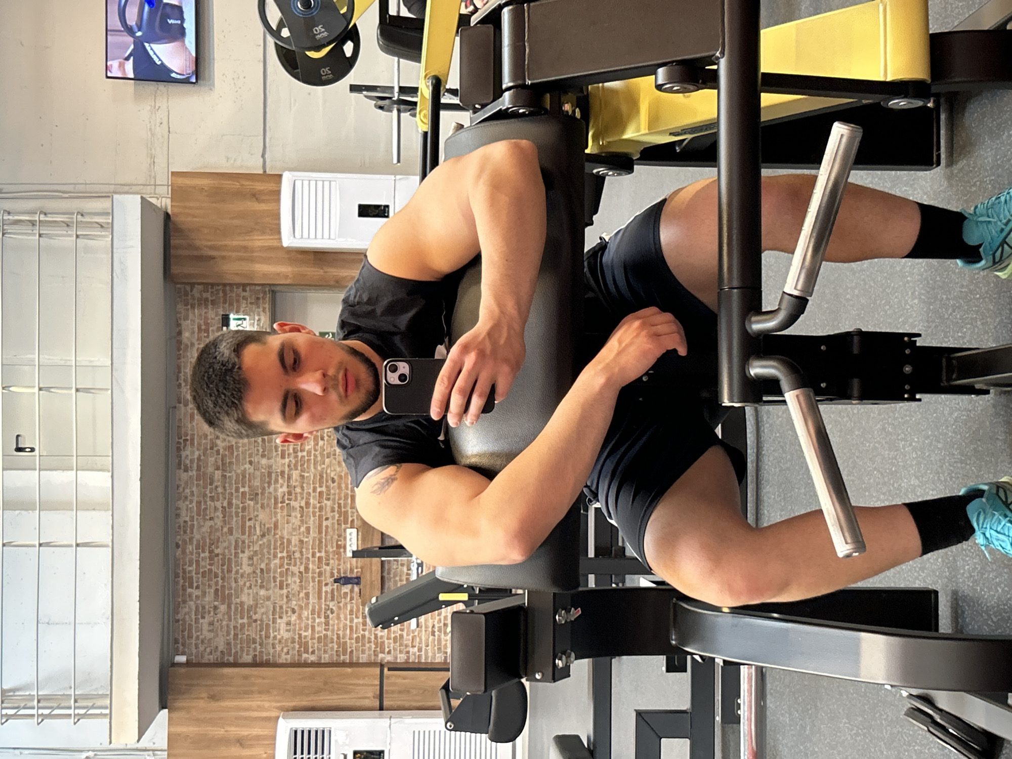 A man is seated on a leg press machine in a gym. He is wearing a black T-shirt, black shorts, and black socks with blue athletic shoes. He is holding a smartphone in his right hand, presumably taking a selfie. The gym features weightlifting equipment, including weight plates, and a television screen is visible in the background. The wall is brick. An air conditioner is visible. There is no verifiable information to confirm the location as Sofia, Bulgaria.