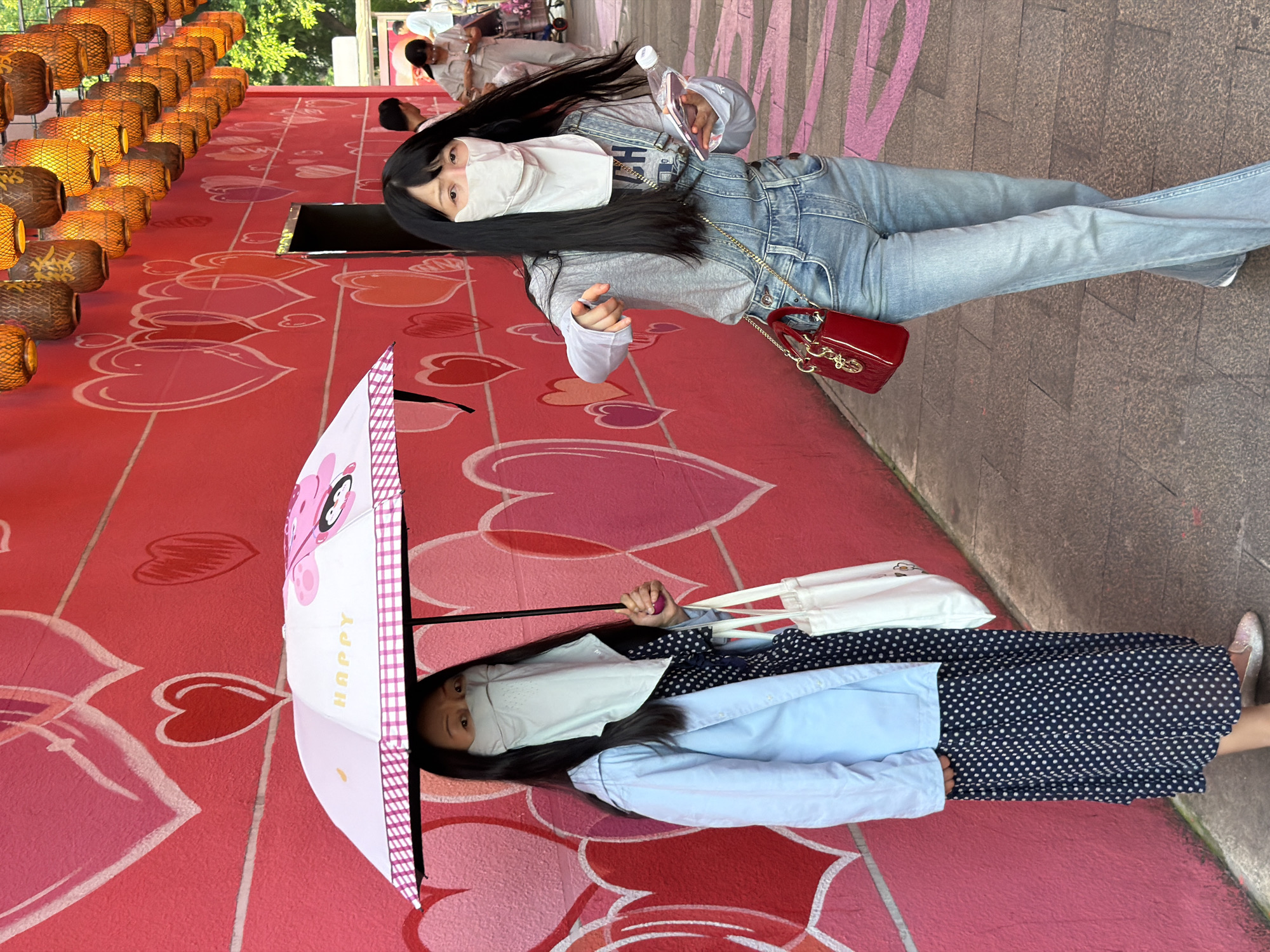 Two women are walking on a sidewalk in Yuzhong District, China, with a bright pink wall adorned with white and pink heart outlines behind them. The woman on the left is holding a pink and white umbrella with a cartoon penguin on it, and she is wearing a light blue jacket over a dark blue and white polka dot dress, a face mask, and silver shoes. She is also carrying a white tote bag. The woman on the right is wearing denim overalls over a light gray long-sleeved shirt, a face mask, and holding a clear plastic bottle and a smartphone. She is also carrying a small red handbag. In the background, several rows of hanging straw lanterns are visible, and sunlight is filtering through trees on the right side of the image. Other people are walking in the distance.