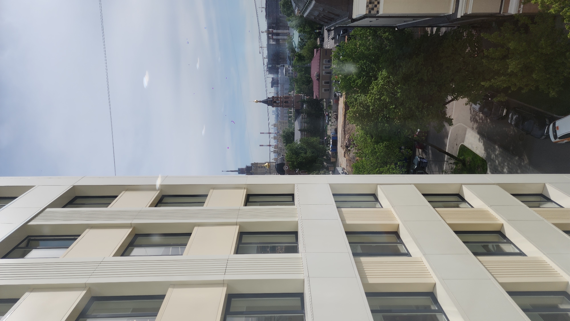 The image captures a cityscape view, likely taken from an upper floor window looking down and across. In the immediate foreground, to the left, is a modern building facade constructed with alternating white and light beige panels, featuring dark-framed windows arranged in a distinct pattern with both horizontal and slanted elements.
Beyond this foreground building, a sprawling urban landscape unfolds under a bright, clear blue sky with some faint clouds. Thin power lines traverse the upper portion of the sky. Numerous green trees are visible throughout the city, indicating spring or summer foliage.
In the mid-distance, several buildings of varying styles can be seen, including older brick structures and more modern multi-story constructions, some of which appear to be under development with cranes visible. A prominent golden-domed Orthodox church stands out, its traditional architecture contrasting with the surrounding city. Further in the background, a tall, dark building with a distinctive spire and golden elements, characteristic of Moscow's Stalinist skyscrapers (one of the "Seven Sisters"), rises above the other structures. Industrial chimneys are also visible on the horizon.
Below the distant cityscape, a paved road or street is visible with several parked cars, including a white van. Some areas appear to be undergoing ground excavation or construction. The day is bright and clear, suggesting late morning or afternoon. There are several reflections and possibly dust specks on the window glass through which the photograph was taken. No legible text is visible in the image. The architectural landmarks confirm the location as Moscow, Russia.