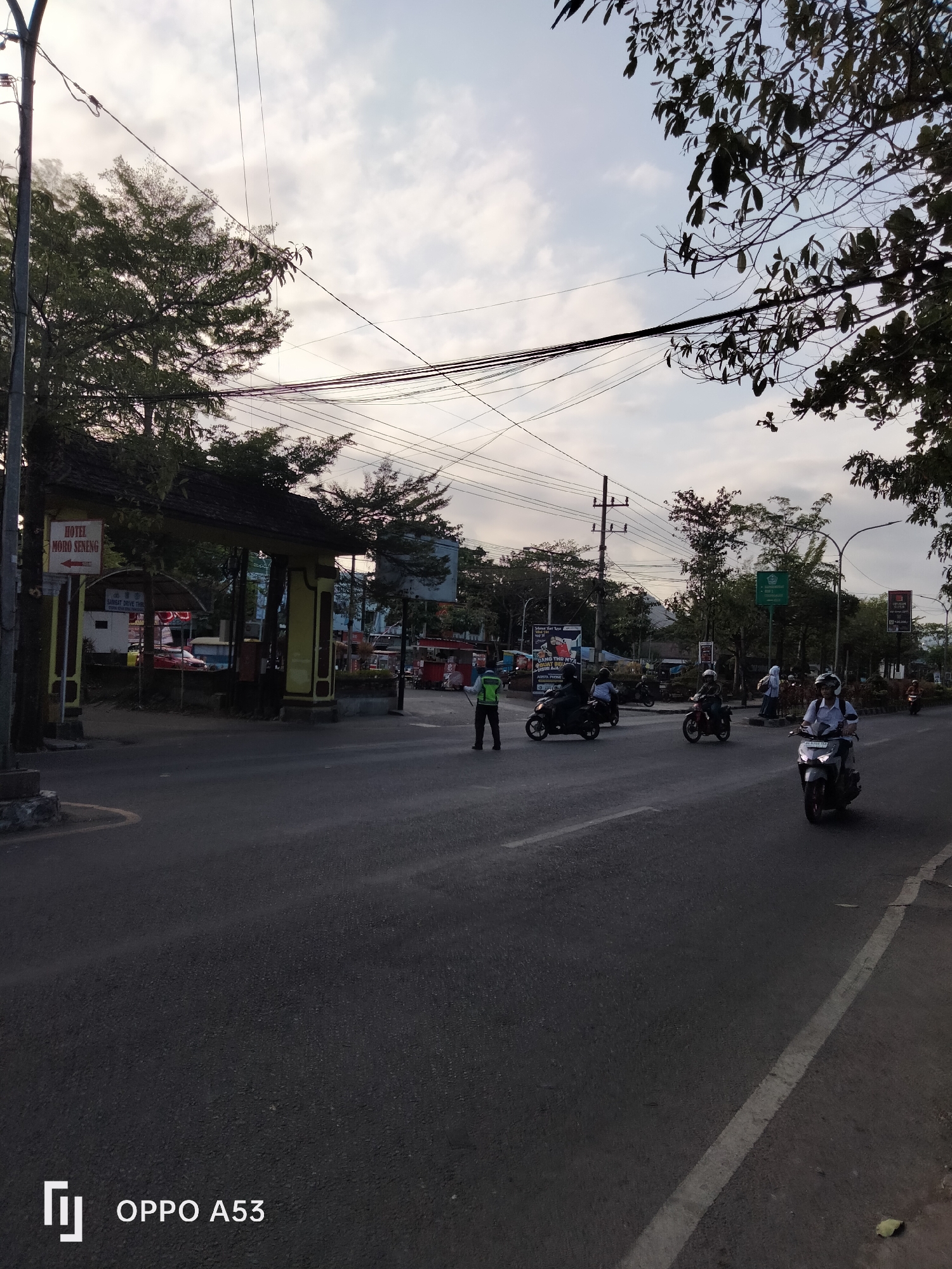 The image depicts a street scene in Kelutan, Indonesia, likely during the late afternoon or early evening due to the soft lighting and visible sky with some clouds. The foreground is dominated by a paved road with a white lane marking. On the left side of the road, a gateway structure with a wooden, dark-tiled roof is visible, featuring a sign that reads "HOTEL MORO SENENG" with an arrow pointing to the left. Next to the gateway, a traffic control officer in a high-visibility vest stands on the road.
Several people are operating motorcycles and scooters on the road. One motorcycle is in the immediate right foreground, with a rider wearing a helmet. Further down the road, other motorcycles are also in motion, carrying one or two occupants. On the right side of the road, beyond the motorcycles, is a sidewalk and trees. In the background, there are buildings, more trees, and utility poles with numerous overhead wires. The overall atmosphere suggests a busy urban or semi-urban environment with ongoing traffic.