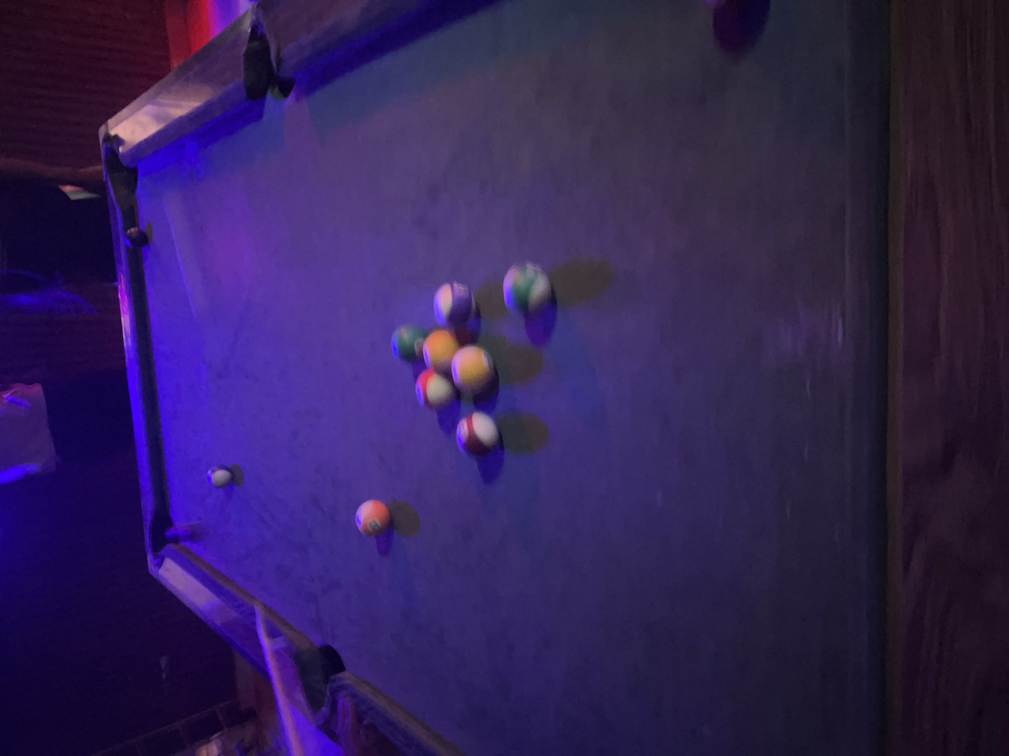 A dimly lit, indoor pool table with several billiard balls scattered across its surface is the subject of this image. The pool table is covered in a purple felt and illuminated by purple and blue lighting, casting shadows. Among the visible balls are an orange and white striped ball, a yellow and white striped ball, a solid red ball, a green and white striped ball, and a solid purple ball. The balls are not arranged in any particular formation, suggesting they may have been recently played. The environment appears to be a recreational space, possibly a bar or game room, indicated by the dark, textured background that resembles wood paneling. There are no people clearly visible in the frame, though a hand and arm are partially visible reaching from the left edge of the image towards the table, suggesting someone is playing or about to play pool. There is no information to suggest the location is in the city of Durimi, Nigeria.