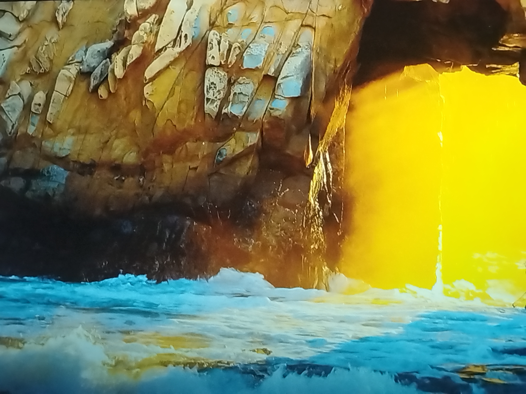 The image depicts a natural scene with a rocky cave opening illuminated by bright yellow light, possibly sunlight. The cave walls are textured and appear to be made of rock with various shades of brown and grey. Some patches of light blue are visible on the rock faces. In the foreground, there is turbulent water with white foam, suggesting waves or a rapid current. The water is a mix of blue and white, with hints of yellow reflecting from the light source. The overall scene suggests an outdoor location, perhaps a coastal area with caves. There are no people, objects, or visible text in the image. The bright light could indicate it is daytime, and the presence of waves suggests a dynamic water environment. The location context of Balashikha, Russia, does not provide specific visual cues that are directly represented in this image, which appears to be a generic natural landscape.