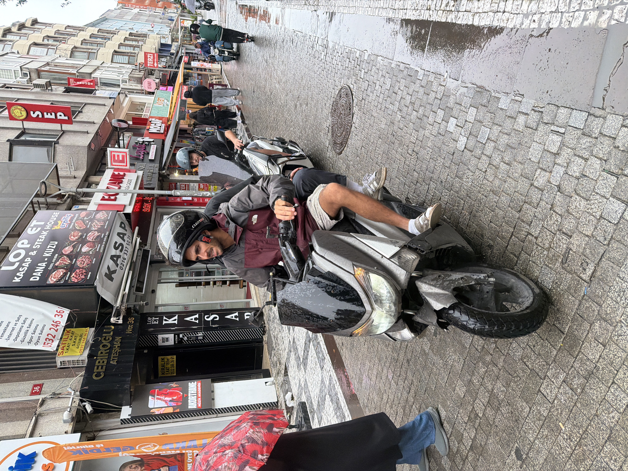A man wearing a maroon and grey jacket, beige shorts, and white sneakers rides a silver scooter with another person behind him. The scooter is positioned on a wet cobblestone street. The man is looking to his right, and his face is visible, showing a slight smile. The street is lined with shops and buildings. One building has a sign that reads "Lop Et Kasap & Steak House". Another sign says "CEBIROĞLU ATEŞHAN". The weather appears to be overcast, and the street is wet, indicating recent rain or current dampness. A person holding a red and black umbrella is partially visible on the left side of the image. In the background, other people are walking, and a few more scooters and motorcycles are parked. The location is identified as Kadıköy, Turkey, specifically on a street with multiple businesses.