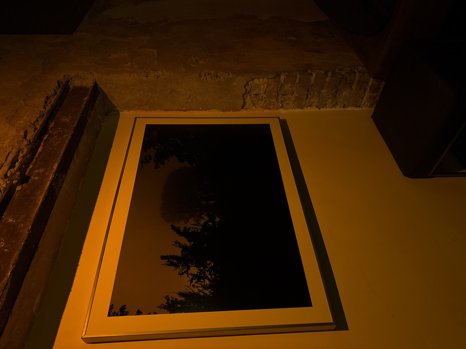 The scene depicts an indoor environment in Sofia, Bulgaria, illuminated by a warm, amber-toned light source. A large, rectangular artwork, oriented horizontally with a white frame, is mounted on a smooth, light-colored wall. The artwork itself features a dark, silhouetted landscape, appearing as dense foliage or trees against a slightly lighter, indistinct background. Above the artwork, a rough, dark, horizontal beam, consistent with timber or a structural lintel, is visible. To the right of the artwork, the wall surface transitions to exposed reddish-brown brickwork, exhibiting signs of missing plaster or render. In the lower right quadrant of the image, a dark-colored, upholstered furniture item, possibly a stool or part of a bench, is partially visible, featuring a minimalist metal frame at its base. No individuals are present.