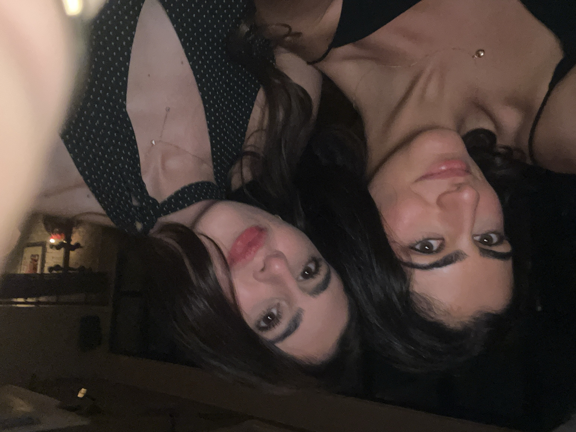 Two women are lying side-by-side, taking a selfie. The woman on the left wears a black and white polka dot dress and red lipstick. The woman on the right wears a black top. Each has dark hair and brown eyes. Both wear necklaces. The background is dark and indistinct, but a decorative shelf with framed art and sculptural items are visible. There is no information to determine if the photo was taken in Paris, France.