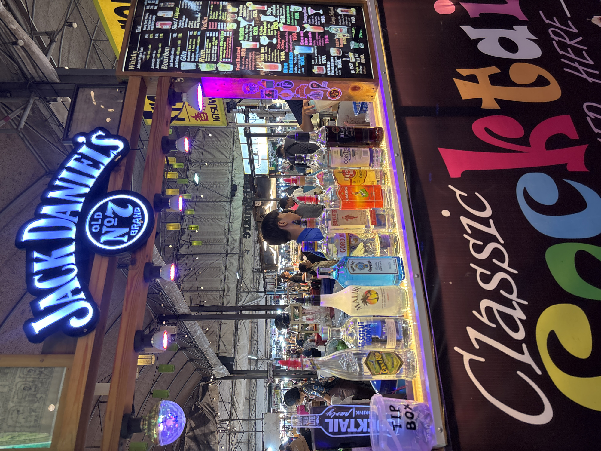 The image depicts an outdoor bar stall at a night market in Chiang Mai City Municipality, Thailand. The stall features a prominent "Jack Daniel's Old No. 7 Brand" neon sign. On the bar counter, a variety of liquor bottles are displayed, including Canario, Absolut Vodka, Malibu, Gordon's, and Bombay Sapphire. A "TIP BOX" is visible on the left. Behind the bar, a menu board lists various cocktails and drinks with prices. Several individuals, including customers and staff, are present in the background, browsing or being served. The environment is dimly lit, with string lights and colored lamps illuminating the market. The overall scene suggests a lively atmosphere typical of a Thai night market.
