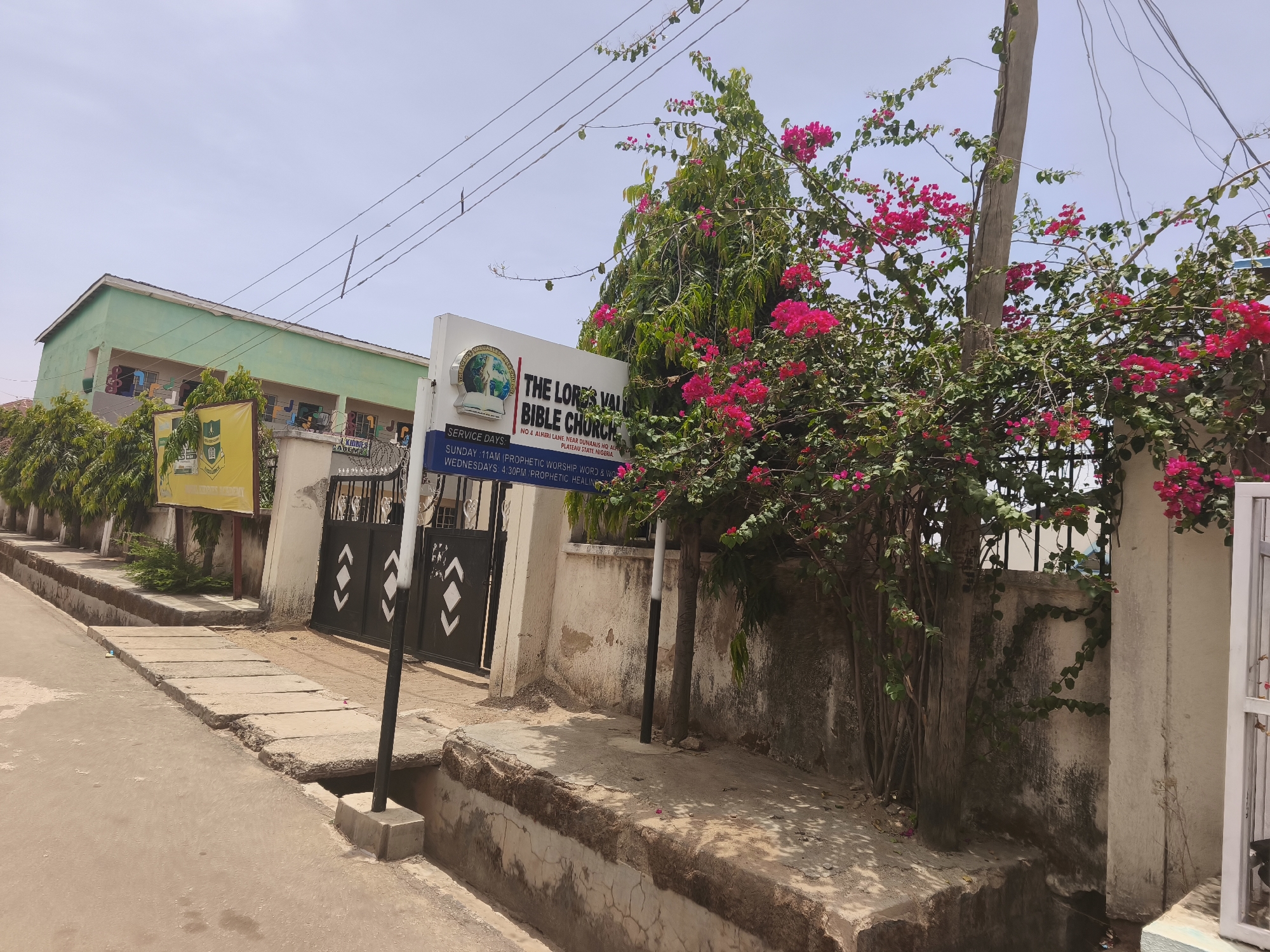 This outdoor daytime photograph captures a street scene in Kabong, Plateau State, Nigeria, featuring the entrance to "The Lords Valley Bible Church" and a school. The weather is clear and sunny, indicated by the bright light and a pale blue sky crisscrossed by utility wires.
In the foreground, an asphalt road runs alongside a concrete sidewalk and a covered drainage system. To the right, a prominent white signboard displays "THE LORDS VALLEY BIBLE CHURCH" with its circular logo. Below the church name, service details are listed: "SERVICE DAYS: SUNDAY: 11AM (PROPHETIC WORSHIP WORD & WO)" and "WEDNESDAYS: 4:30PM PROPHETIC HEALING". The address "NO 4 ALHERI LANE, NEAR DUNAMIS HQ ALFRE, PLATEAU STATE, NIGERIA" confirms the location.
Behind the sign and to its left, a black metal gate with distinctive white arrow-like patterns marks the entrance to the property. Lush green foliage, including a large bush heavily adorned with vibrant pink bougainvillea flowers, frames the right side of the entrance and extends towards the gate. A tree trunk is visible amidst the flowers.
In the background on the left, a light green building with a dark roof and multiple windows can be seen. A yellow banner or sign, partially visible in front of this building, reads "KIDDIES ACADEMY," suggesting a school is part of the complex. No people are visible, presenting a quiet, bright scene under the clear Nigerian sky.