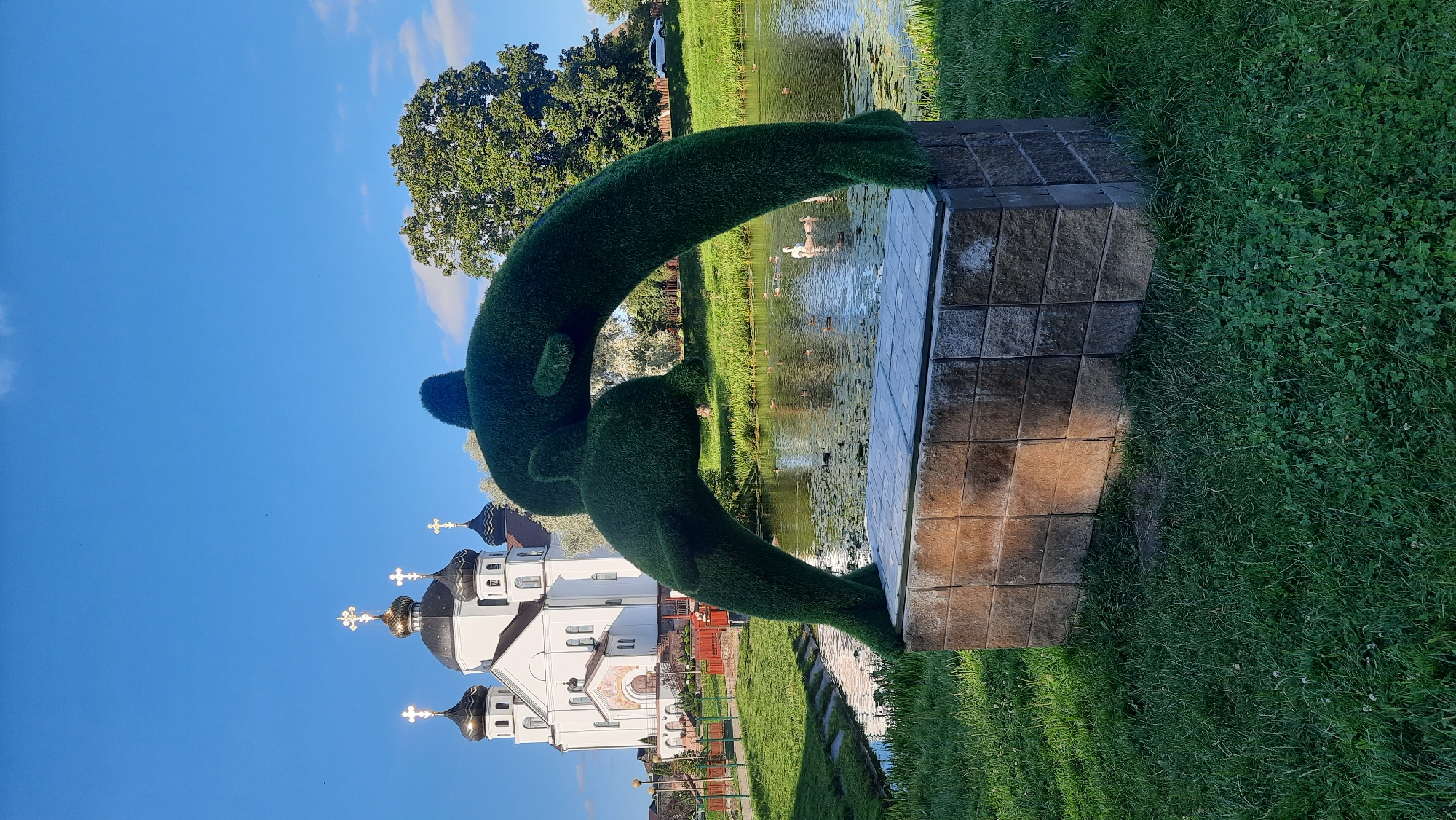 This image depicts a sunlit outdoor scene at the Svyato-Uspensky Convent in Smarhon, Belarus. In the foreground, a topiary sculpture of two dolphins, crafted from dark green artificial turf, is perched on a rectangular stone pedestal. The pedestal is constructed from stacked, rectangular stone blocks with a weathered, rough texture, predominantly in shades of brown and grey. The sculpture faces a tranquil body of water, likely a pond or canal, where several ducks and two white swans are swimming amongst lily pads.
In the middle ground, a meticulously maintained green lawn surrounds the pond and sculpture. Beyond the pond, the white facade of the Svyato-Uspensky Convent stands prominently. The convent features multiple domes topped with golden crosses, characteristic of Orthodox Christian architecture. The sky above is a clear, vibrant blue with a few wispy white clouds. To the right of the convent, a large, mature green tree provides a backdrop, and a white car is visible on a paved area in the distance. The overall atmosphere is serene and peaceful, bathed in the warm light of what appears to be late afternoon.