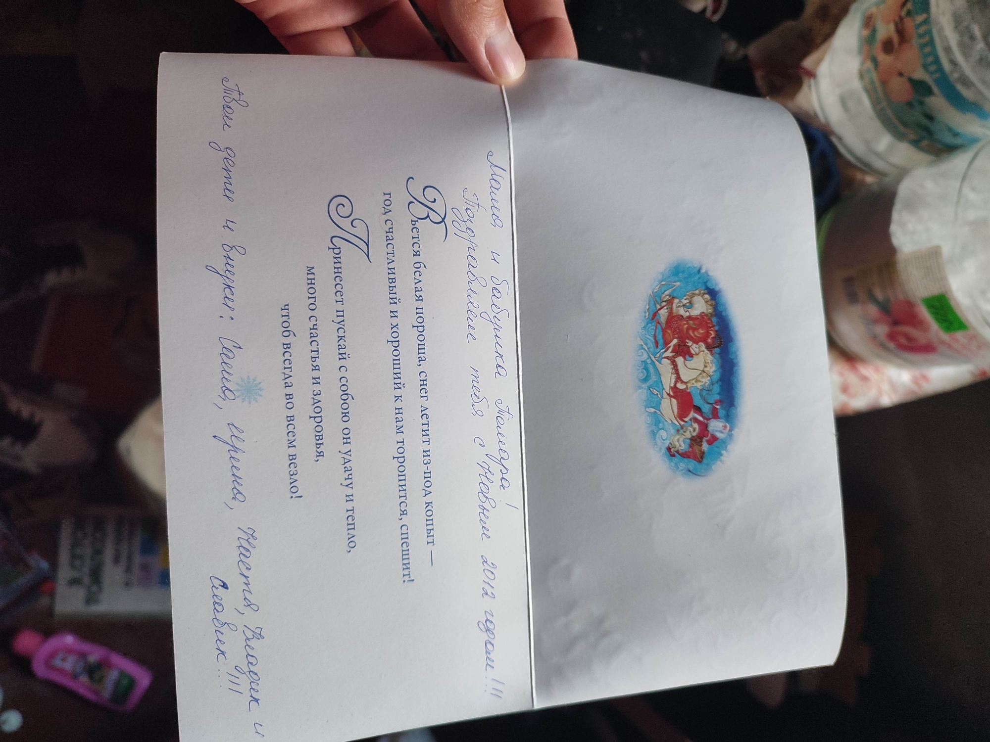 The image shows a handwritten greeting card, likely a Christmas or New Year's card, with a festive illustration of Santa Claus. The card is held by a person's hands, suggesting it is being presented or read. The background is dark and appears to be an indoor setting, with some out-of-focus objects like jars and a pink bottle visible.
The card itself contains a handwritten message in Russian. The top portion of the card features a decorative illustration depicting Santa Claus riding a sleigh pulled by reindeer, set against a blue and white swirling background.
The handwritten text on the card, translated from Russian, reads:
"Mom and Grandma Pomara!
Congratulations
Wishing you a Happy New Year 2012!!!
May the white snow swirling from under the hooves,
The happy and good year hurries to us, rushes!
May it bring you luck and warmth,
Much happiness and health,
So that everything goes well in everything!
Your children and grandchildren: Sasha, Serina, Nastya, Vladik"
The text expresses warm wishes and congratulations for the New Year, specifically for "Mom" and "Grandma Pomara". The mention of "New Year 2012" indicates the time frame of the greeting. The cursive handwriting and the festive imagery suggest a personal and heartfelt message. The overall tone of the card is joyful and affectionate.