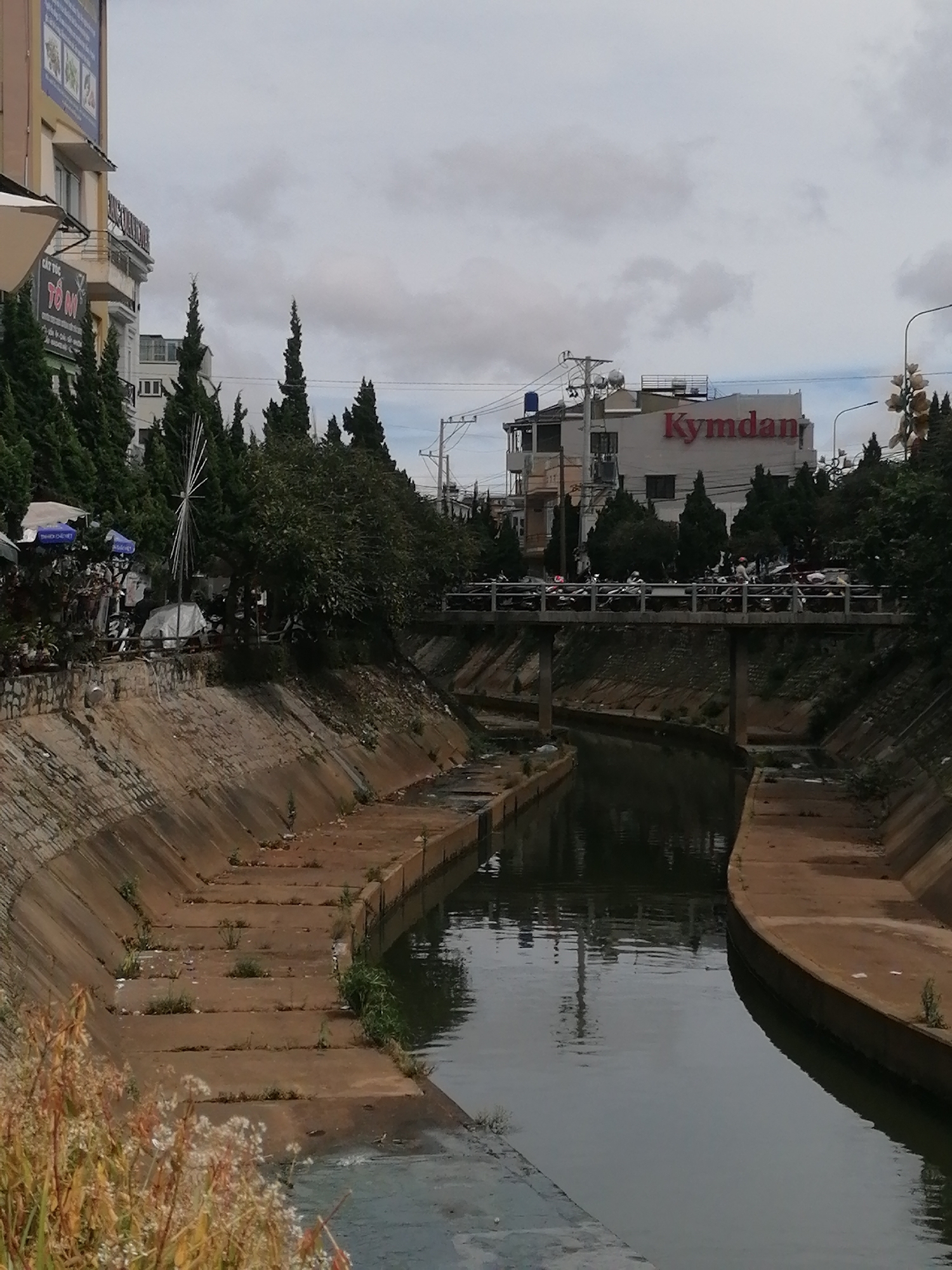 The image shows a concrete canal running through an urban area in Da Lat, Vietnam. The canal has steep, sloped sides lined with concrete. The water in the canal is dark and reflects the overcast sky and surrounding buildings. On the left side of the canal, there are buildings with signs, one of which appears to be a shop with an awning. A series of tiered concrete steps lead down to the canal on the left. On the right side of the canal, the concrete embankment continues. A bridge crosses the canal in the middle of the image, with many motorcycles parked on it. Buildings are visible on either side of the canal, including a building with a red sign that reads "Kymdan". The sky is cloudy, suggesting it is an overcast day. Several tall, thin evergreen trees are planted along the banks of the canal and near the buildings. In the foreground on the left, there is dry, golden vegetation. The scene appears to be a commercial or residential area within the city.