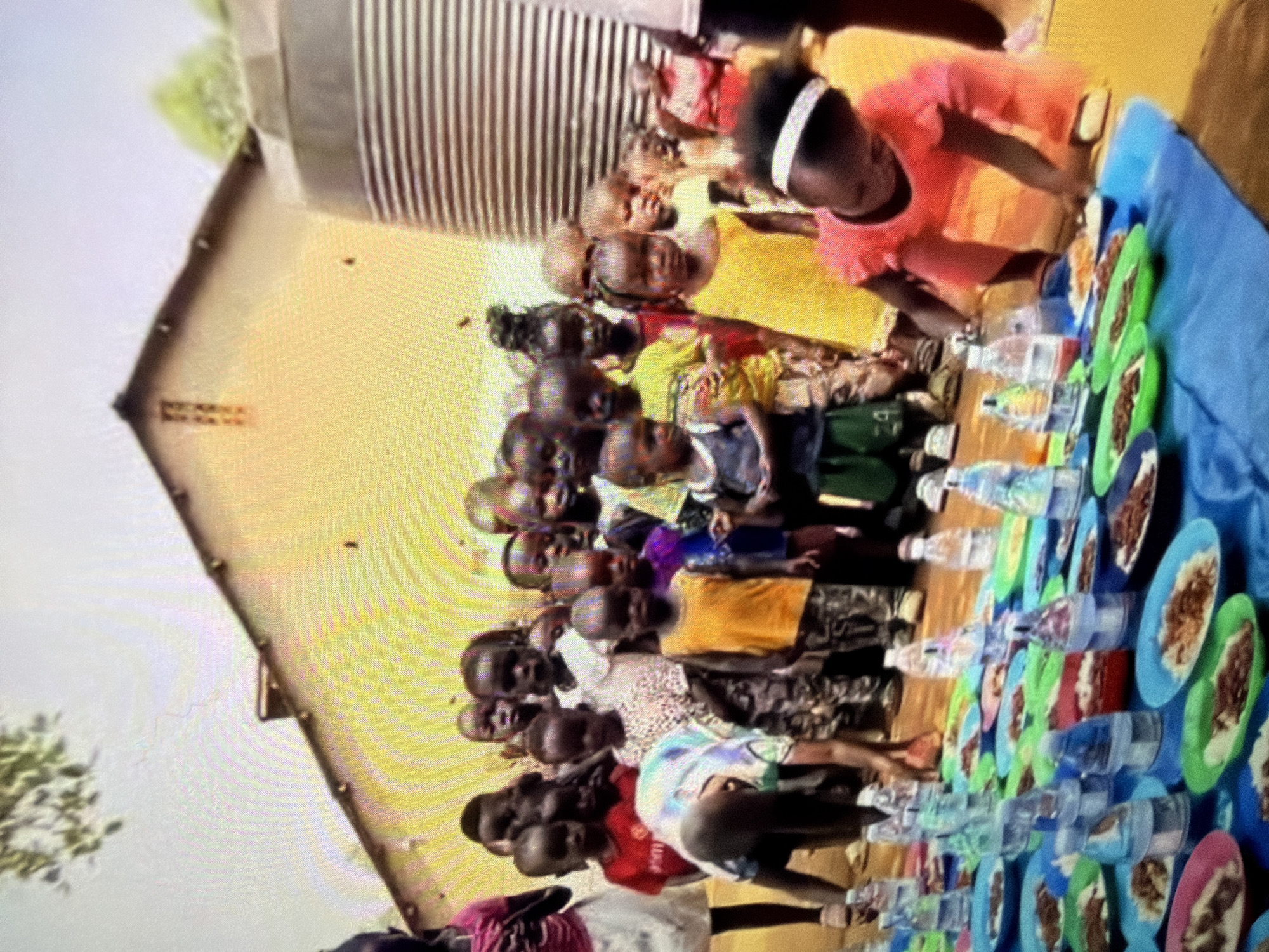 A group of children are gathered around a table covered with a blue cloth. On the table, there are many plates of food and bottles of water, suggesting a meal is about to be served or is in progress. The children are of various ages and appear to be in an outdoor setting, with a building and a large water tank visible in the background. The children are mostly looking towards the camera or interacting with the food. The overall scene suggests a communal meal or a charitable event for children.