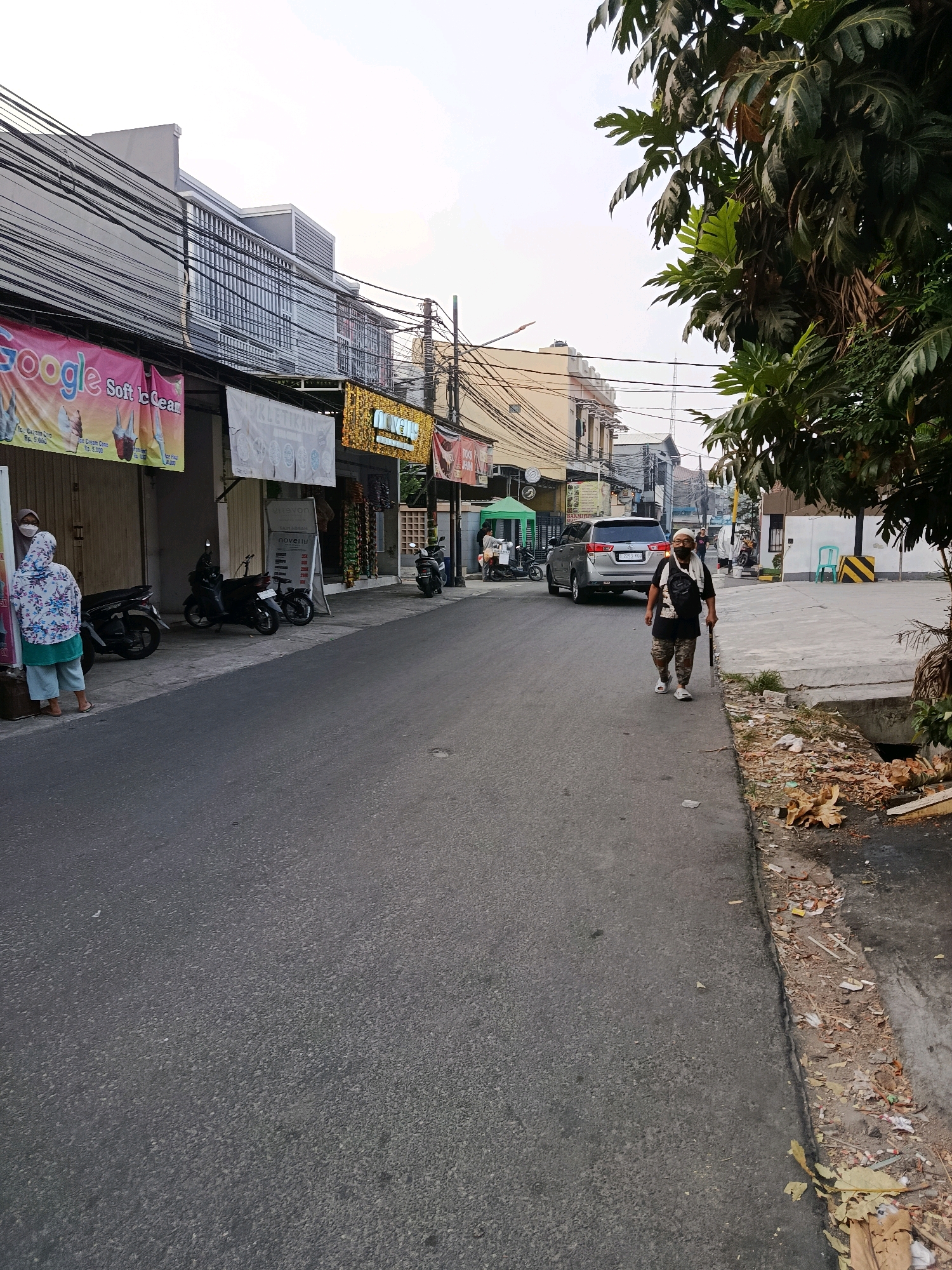 In Bekasi, Indonesia, on a paved road in an urban setting, a lone individual walks from right to left in the mid-ground. This person, a man, wears a white head covering, a dark t-shirt, camouflage-patterned shorts, and white athletic shoes. He carries a dark bag over his left shoulder and a dark walking stick in his right hand. The man is positioned on the right side of the road, close to the sidewalk, which is littered with debris, including dried leaves and trash.
To the left of the road, a silver SUV is parked. Further left, several motorcycles are parked along the sidewalk in front of businesses. On the far left, a woman in a floral-patterned headscarf and a light blue-green patterned tunic stands near a motorcycle, facing towards the street. Above the businesses, a dense network of electrical wires crisscrosses overhead.
The buildings lining the street are a mix of commercial establishments with awnings and signs. One prominent banner displays the word "Google" and "Soft Ice Cream" in colorful lettering. Another sign shows "NOVETIS". The environment appears to be a commercial street with moderate activity. The sky is overcast.