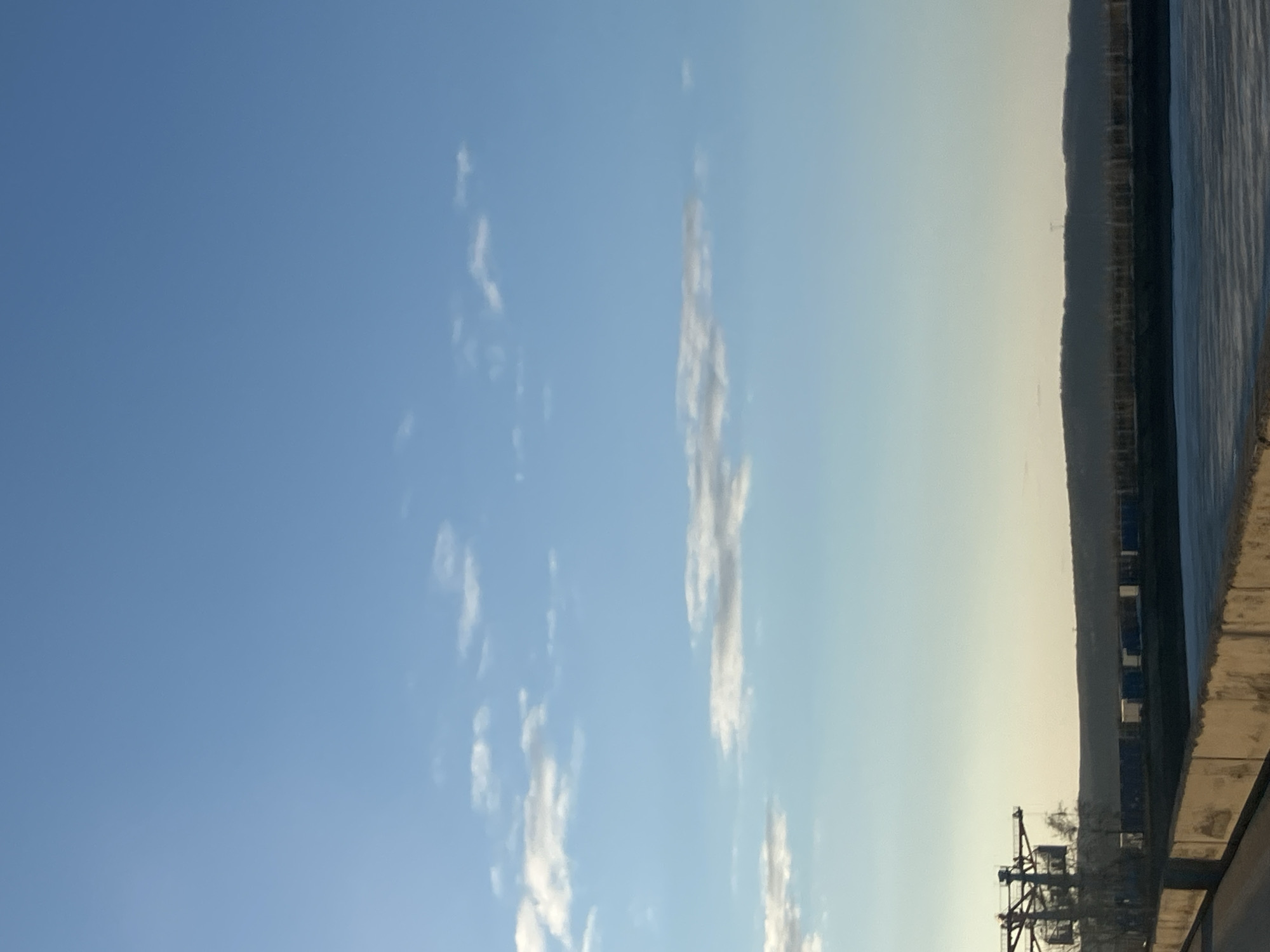 The image depicts a view of the Kama River from what appears to be a riverside promenade in Chaikovsky, Russia. The sky is predominantly blue with scattered cirrus clouds. A land mass (possibly an island or the opposite bank) is visible across the river, featuring trees along the shoreline and what may be utility poles. In the foreground, a concrete barrier or wall is visible. To the right of it is the bank side, where machinery and cargo container storage facilities are located. The overall scene suggests a location along the Kama River in Chaikovsky, with industrial activity adjacent to the river's edge. The image appears to have been taken during daylight hours.