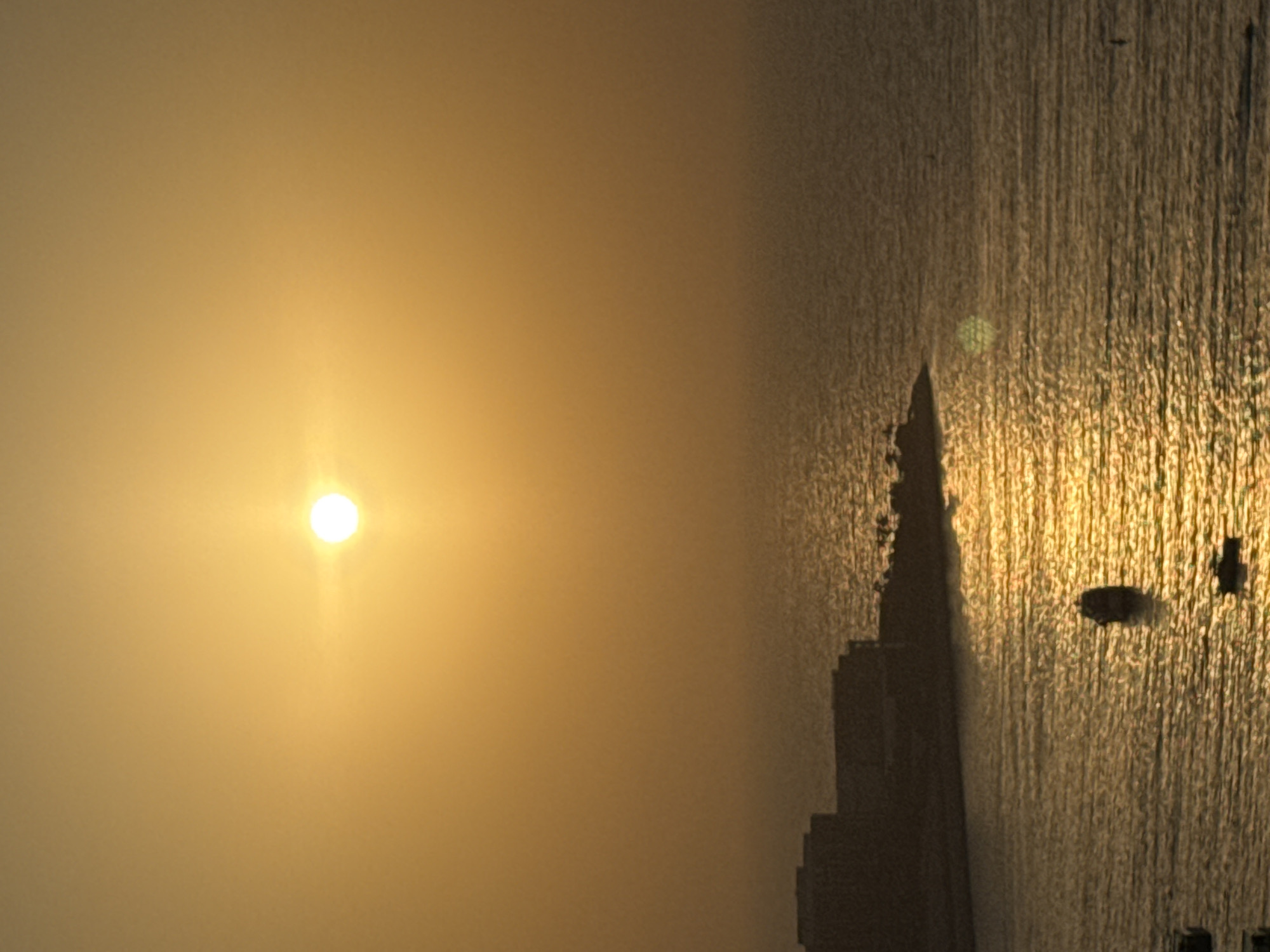This image captures a serene sunset over the waters of Dubai, United Arab Emirates. The sun, a bright, luminous orb, hangs low in a hazy, golden sky, casting a warm glow across the scene. In the foreground, the calm sea reflects the sunset's light, creating shimmering patterns on its surface. Several small boats are visible on the water, adding a touch of human activity to the tranquil setting. In the background, a silhouette of modern buildings lines the coast, suggesting the vibrant cityscape of Dubai. The overall impression is one of peaceful transition from day to night, with the natural beauty of the sunset harmonizing with the urban landscape.