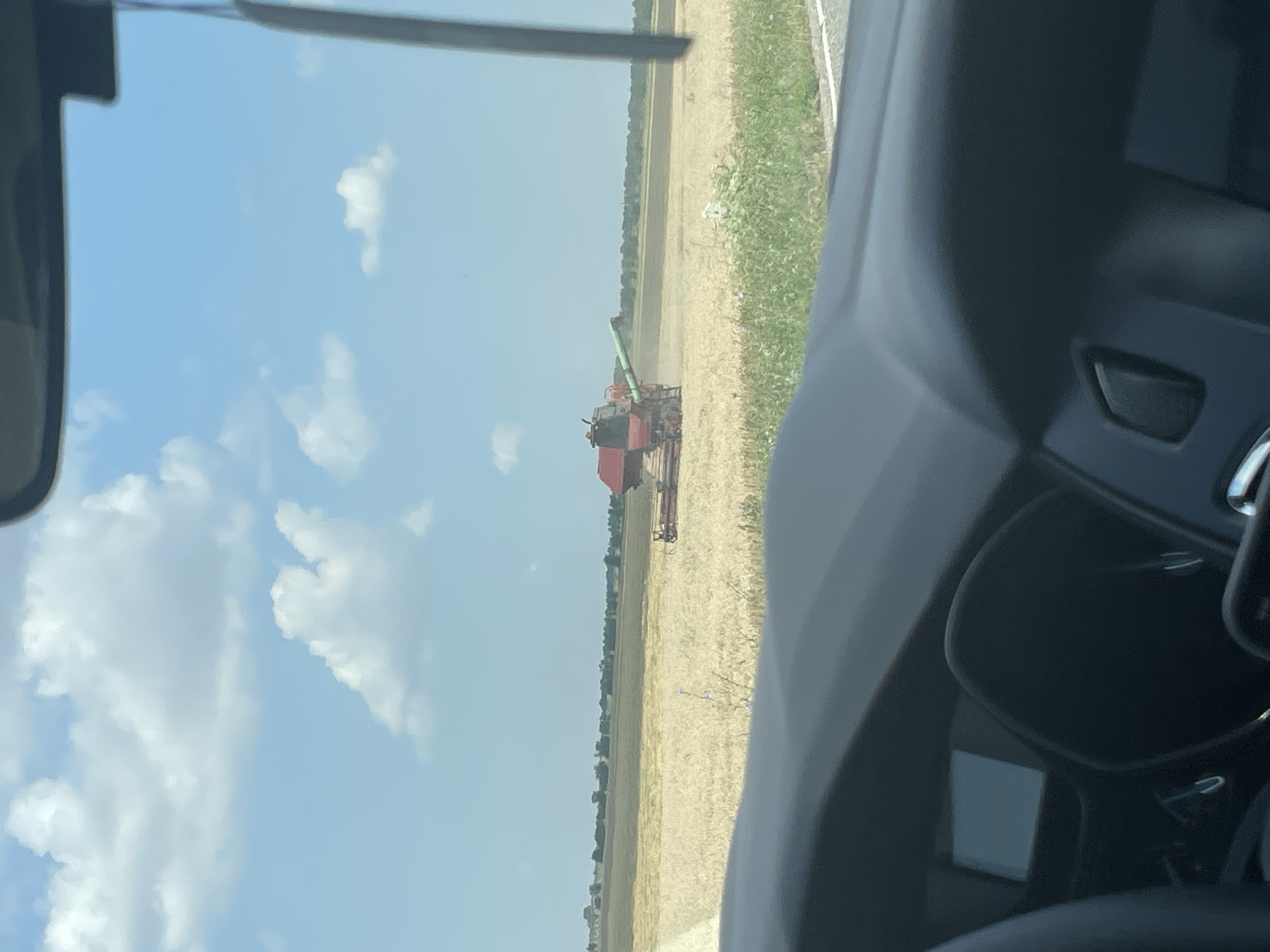 The image shows a combine harvester working in a field of harvested grain under a partly cloudy sky. The combine is red and green and is positioned in the middle-right of the frame. The field is mostly golden with some green vegetation in the foreground. The perspective is from inside a vehicle, with the dashboard visible in the lower part of the image. A rearview mirror is visible in the upper right corner. The location is identified as Proletarskii, Russia.
