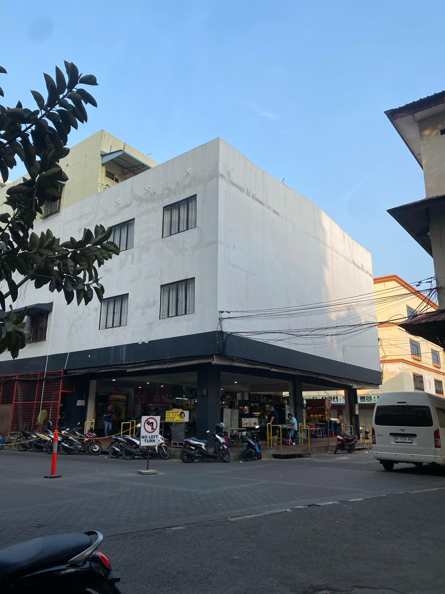 The image shows a street scene in Batam, Indonesia, with a prominent white building on the right side. The building has multiple floors with dark-framed windows. The ground floor appears to be a commercial establishment with an open storefront, displaying goods and several individuals inside. Parked motorcycles are lined up along the sidewalk in front of the building. A white van with license plate BP 7210 DE is parked on the street. A traffic sign indicating "NO LEFT TURN" is visible near the motorcycles. The sky is clear with a gradient from light blue at the top to a warmer tone near the horizon. Trees with dark green leaves are partially visible in the foreground, on the left side of the image. The overall environment suggests a commercial area in a city.