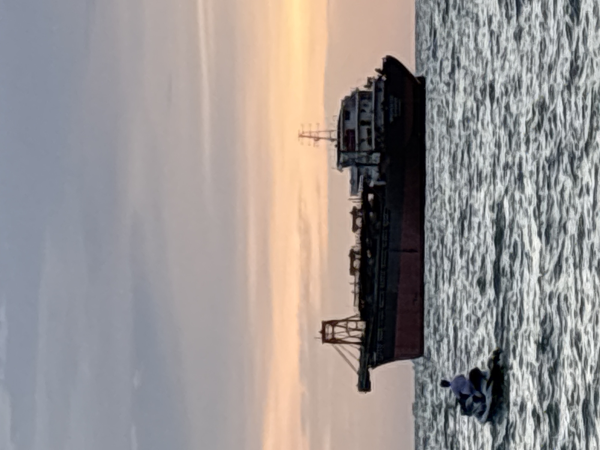A cargo ship is anchored offshore in Pattaya City, Thailand, during sunset. The sky is a mix of grey clouds and warm orange hues from the setting sun. The ship is large and appears to be carrying cargo. In the foreground, a person is riding a jet ski on the choppy water. The water is dark and reflects the colors of the sky. The overall scene depicts a peaceful maritime moment at dusk in Pattaya City.
