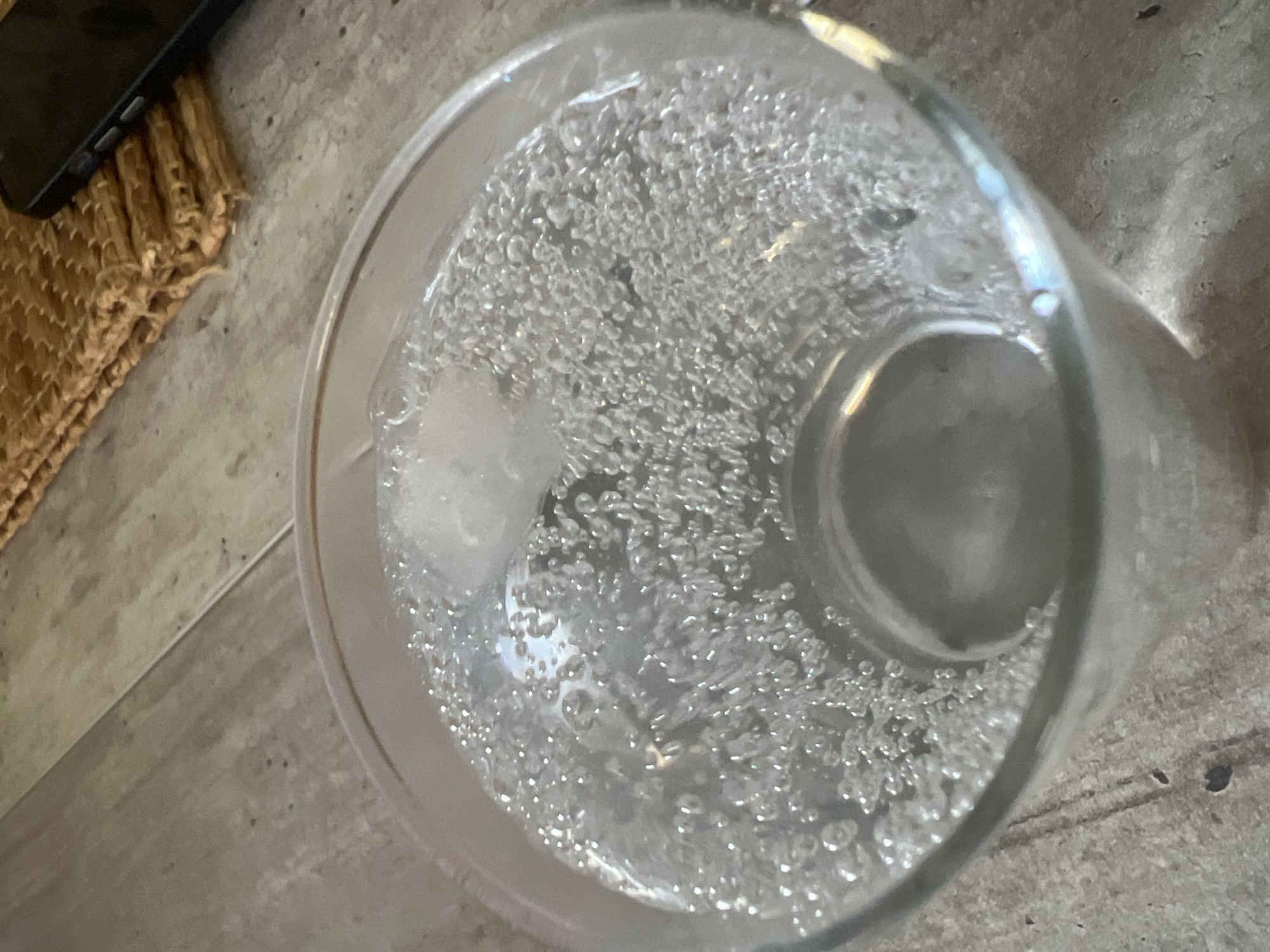 A glass of bubbly water with ice sits on a wooden surface, next to a woven placemat and a smartphone. The image is taken from a high angle, looking down into the glass. The background is blurred, but appears to be an indoor setting. There are no people visible in the image. The scene does not provide any specific clues about its location in Volgograd, Russia.