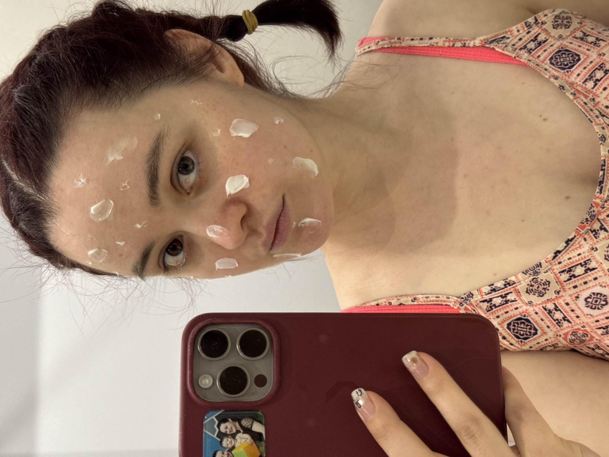 A woman with fair skin and dark auburn hair is holding a burgundy-colored iPhone to take a selfie. Her hair is pulled back in a ponytail with a yellow hair tie. She has dabs of a white cream on her face, specifically on her forehead, cheeks, and around her mouth. The woman is wearing a patterned swimsuit top with orange and beige tones. The iPhone has a sticker of a group of people on the back. The background appears to be a plain, white wall. There is no indication of the location in the city of Izhevsk, Russia.