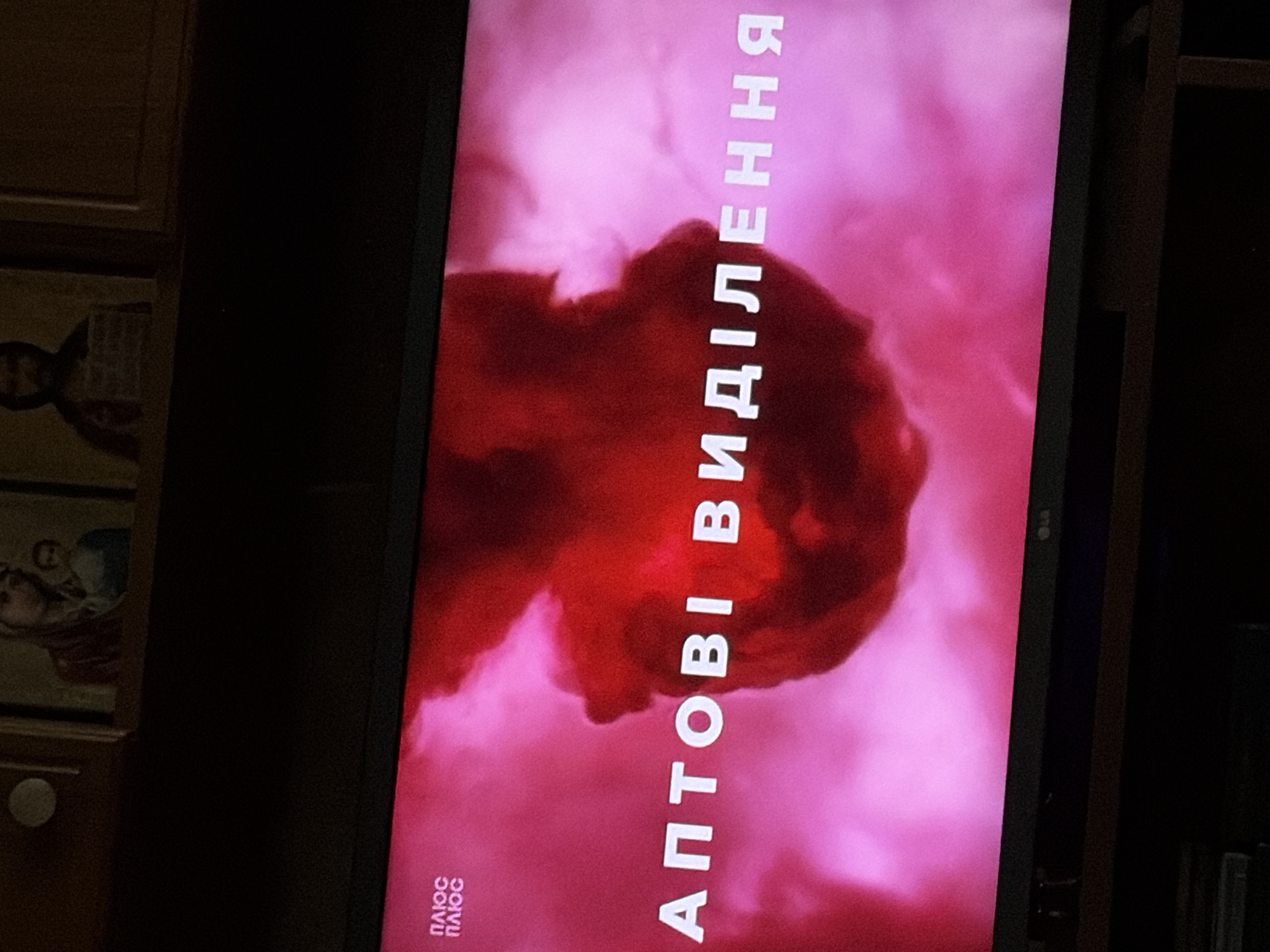 A television screen displays text and imagery against a vibrant pink and red background. In the upper left corner of the screen, the Ukrainian text "ПЛЮС ПЛЮС" is visible in white lettering. The main text on the screen reads "АПТОВІ ВИДІЛЕННЯ" in large white capital letters. The background features abstract, cloud-like formations in shades of red and pink, with a darker, more solid shape in the center. The overall impression is of a graphic display, likely from a television program or advertisement. The scene is set in an indoor environment, with the television placed on a dark piece of furniture. Above the television, two religious icons are partially visible. The resolution of the image is moderate, with some graininess.