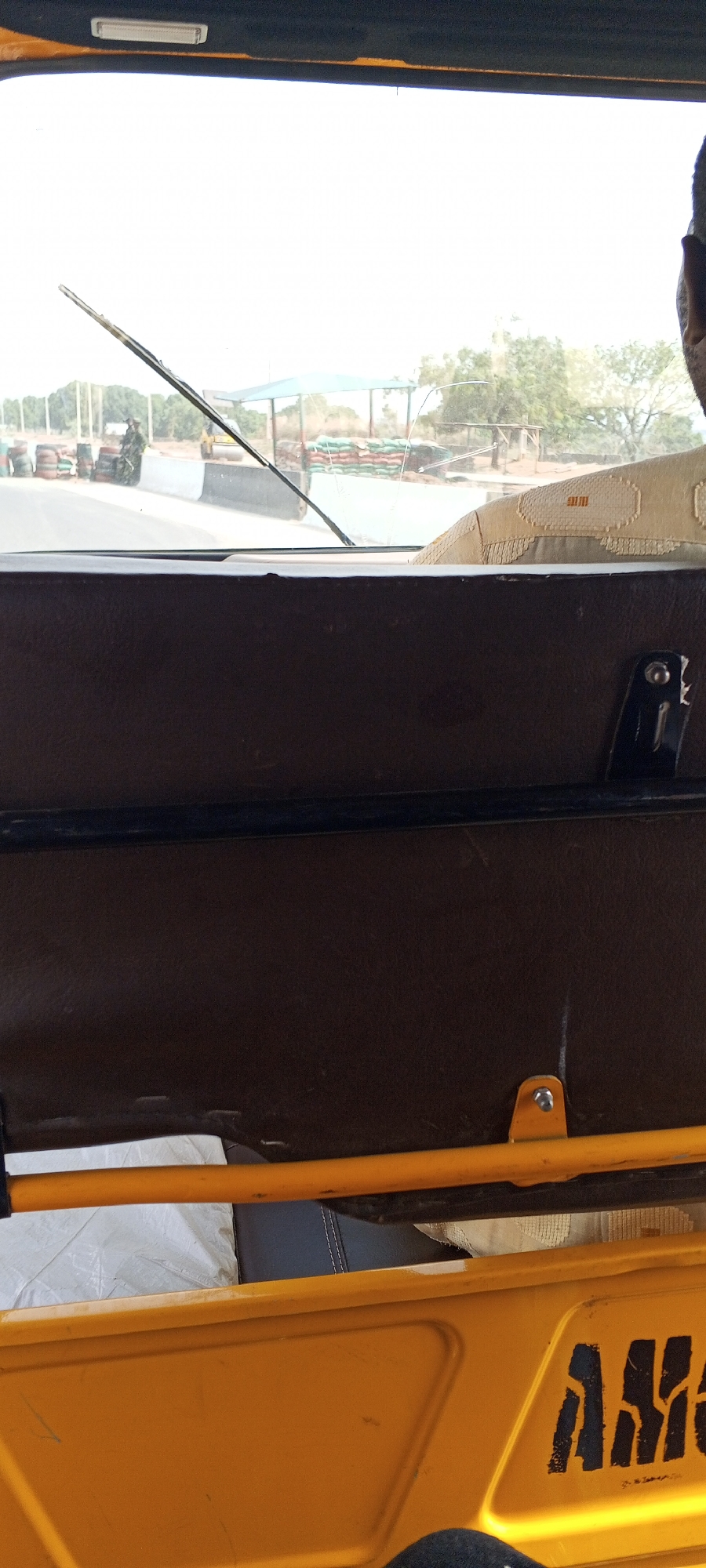 The image shows a view from inside a yellow auto rickshaw, commonly used as transportation in Nigeria. Outside the vehicle, the scene features a military or security checkpoint near Uke, Nigeria. This is indicated by the presence of uniformed personnel, tires forming barriers, sandbags, and a guard post structure. A road roller vehicle is also visible. Inside the rickshaw, the view is partially obstructed by the vehicle's brown interior paneling and structural elements.