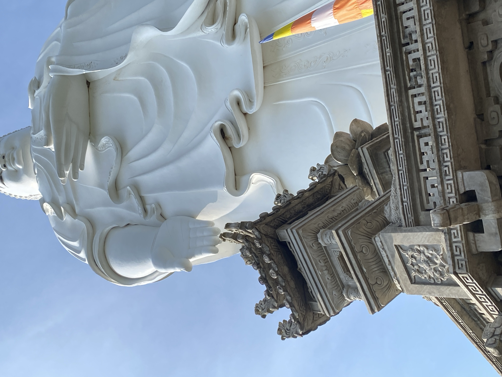 The image shows a low-angle, close-up view of a colossal white statue, likely a Buddhist deity, against a clear blue sky. The statue is predominantly white and features flowing robes. In the foreground, the ornate stone roof of a smaller structure is visible, adorned with intricate carvings and decorative elements. The architectural style suggests an East Asian influence. A vertically striped flag, with colors of orange, white, and blue, is partially visible to the right of the statue, near its base. The scene is set in Tay Nha Trang Ward, Vietnam.