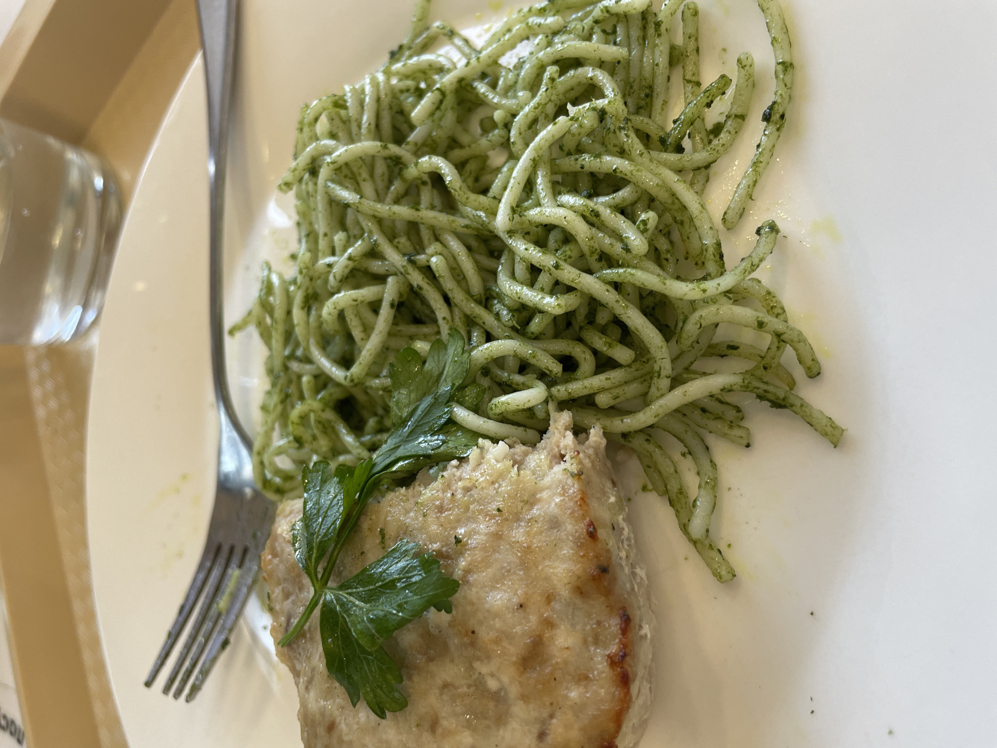 A top-down view shows a white plate containing cooked pasta with a green pesto-like sauce and a piece of cooked meat, garnished with a sprig of parsley. A metal fork is positioned on the left side of the plate. A partial view of a clear glass and a light-colored surface are visible in the background. The location is unknown, as the image does not provide any details about Izhevsk, Russia.