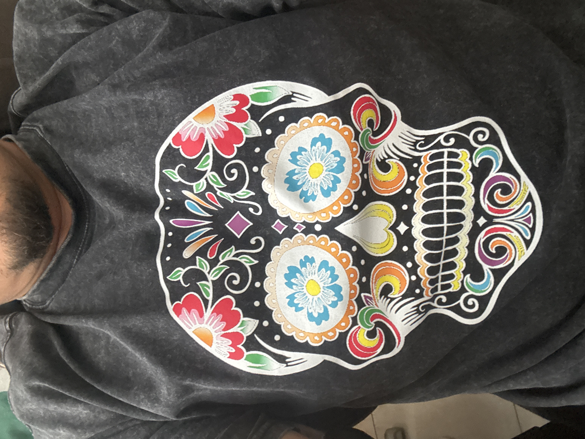 The image shows a person wearing a gray shirt with a colorful sugar skull design on the front. The skull is white with floral and decorative elements in red, orange, yellow, blue, green, and purple. The person's beard is visible. The location is unknown.