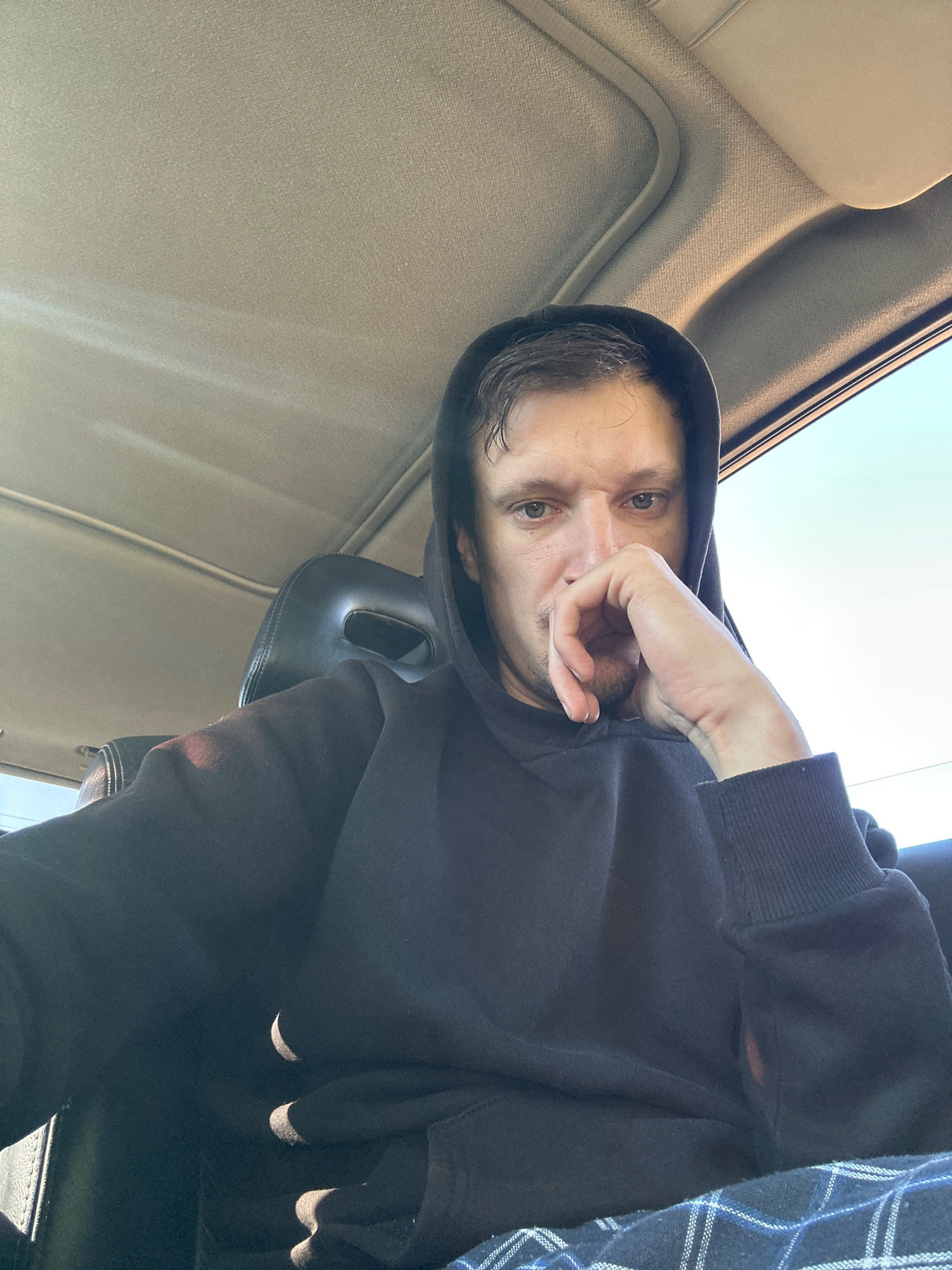 The image shows a selfie of a man wearing a black hoodie, seated in a car. The man is looking towards the camera with his hand resting on his chin. The car's interior is visible, including the headrest and parts of the ceiling. Sunlight streams in from the right side of the car, casting shadows and highlighting the man's face and clothing. The background shows a bright, overcast sky. There is no indication of the location being the city of Makiv, Ukraine.