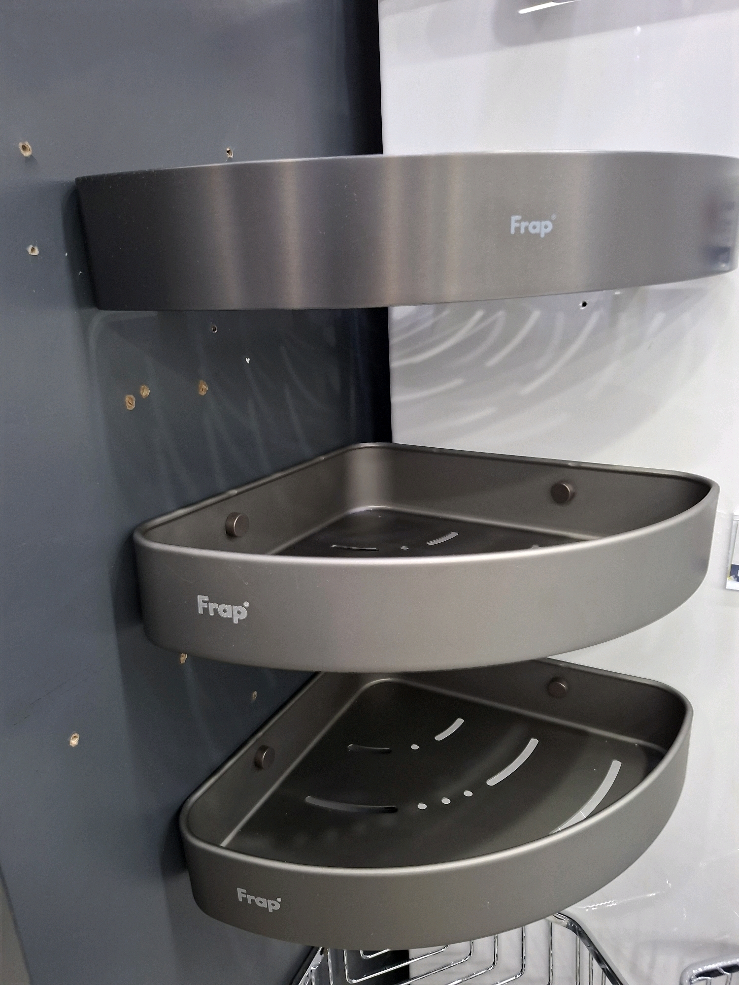 The image displays a close-up, eye-level view of a dark gray corner shelf unit with three tiers, mounted on a wall. Each shelf is triangular and has a raised lip. The top shelf features the "Frap" logo in white lettering on its front edge. The middle shelf also has the "Frap" logo, and a circular metallic fastener is visible on its inner side. The bottom shelf, partially obscured, also bears the "Frap" logo. The wall behind the shelves is primarily a dark gray color with several small, circular imperfections or holes. To the right of the shelves, a white surface is visible, with faint, wavy patterns. The overall setting appears to be an indoor environment, possibly a bathroom or shower area, given the nature of the shelving unit. The image does not provide any discernible information about the city of South Tangerang, Indonesia, or any specific geographical context. There are no people or animals present in the image.
