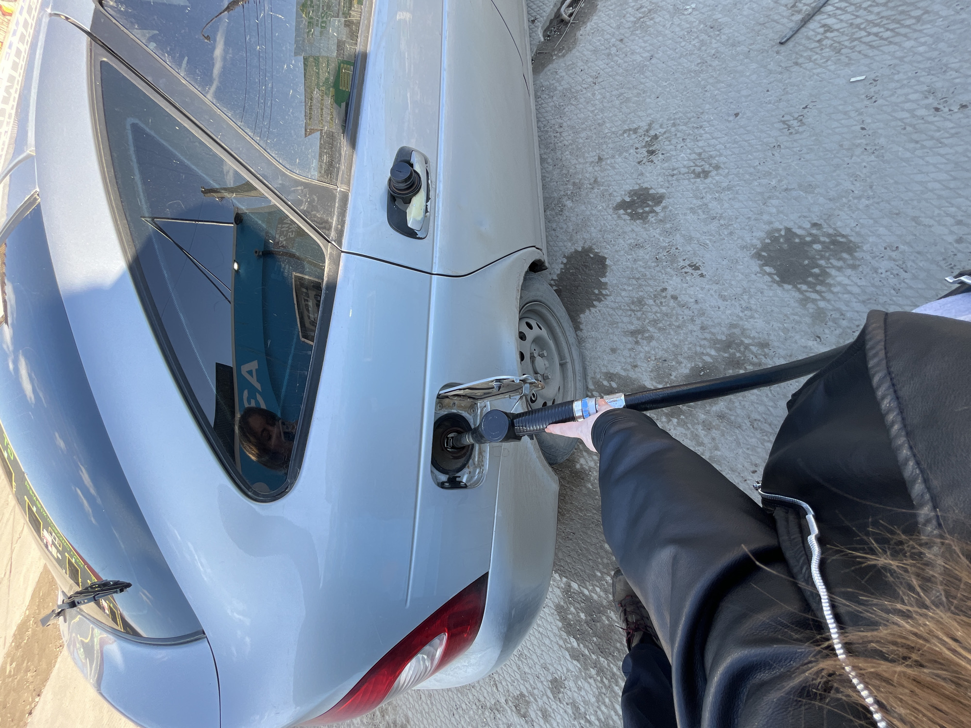 A person is refueling a light gray sedan at a gas station. The car is positioned with its rear facing the viewer. A black fuel pump nozzle is inserted into the fuel tank opening. The person holding the fuel pump is wearing a black leather jacket. The ground is concrete with some discoloration. The car's rear windshield reflects a gas station sign. The image was taken in the city of Chaikovsky, Russia.