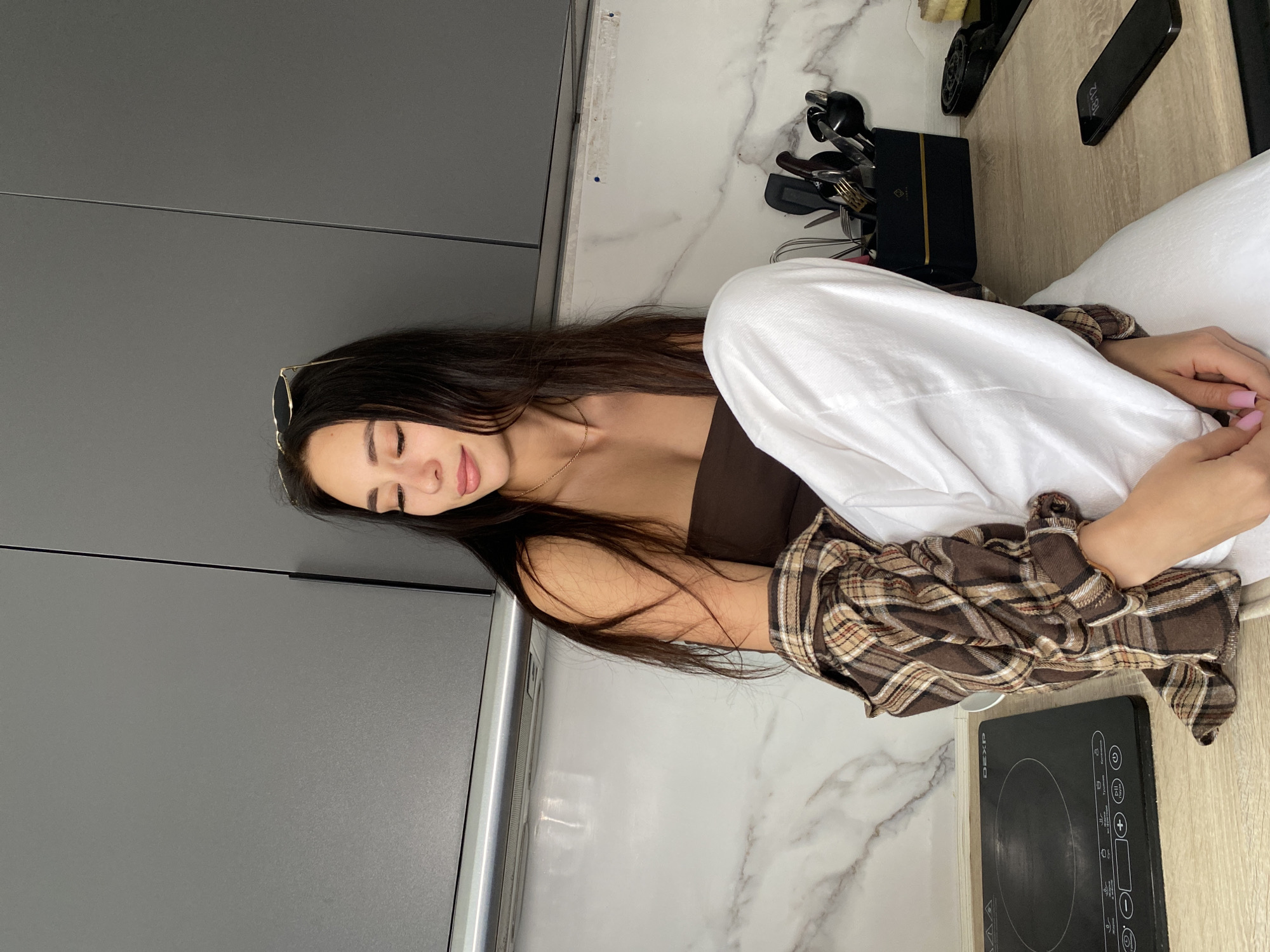 A young woman with long dark hair is sitting on a countertop in what appears to be a kitchen. She is wearing a brown top, white pants, and a brown and tan plaid shirt. She has sunglasses perched on her head. Her eyes are closed. To her right is a black stovetop. Other kitchen items, including a knife block and a cell phone, are visible on the counter. The countertop is made of a white marble-like material, and grey cabinets are visible on the left side of the frame. The location is an indoors scene, likely in the city of Chaikovsky, Russia.