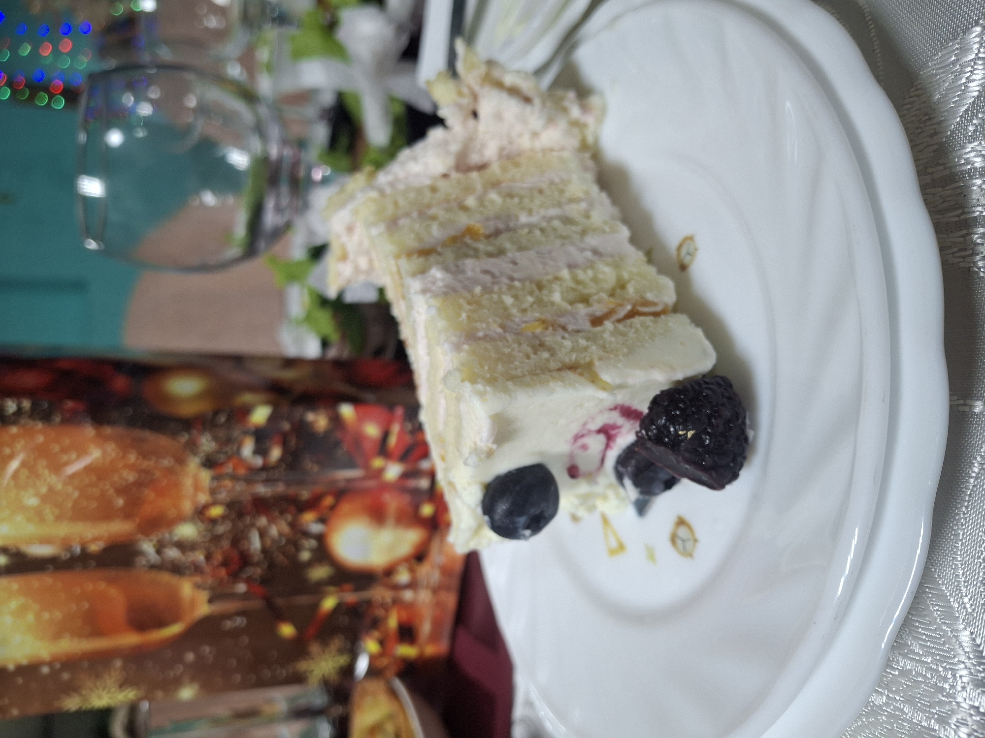 A close-up shot captures a slice of layered cake adorned with blueberries and blackberries, presented on a white plate. The cake features multiple layers of light-colored sponge interspersed with cream filling and topped with a pale frosting. To the left, a blurred scene includes a wine glass and decorative lights, suggesting a festive or celebratory atmosphere. The background reveals a patterned surface, which may be a table covering. It is difficult to confirm the location as Chaikovsky, Russia without additional context or identifying elements within the image.