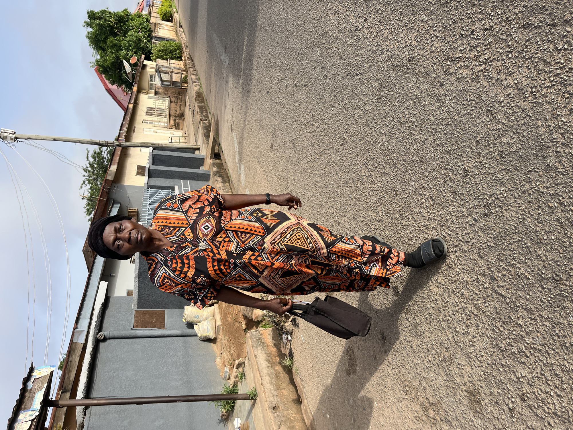 A middle-aged woman stands on a paved street in Jos, Nigeria. She is dressed in a colorful, patterned orange and black dress and black footwear. A black head covering adorns her head. She holds a dark-colored bag in her left hand. Behind her, a mix of buildings line the street, including a house with a red roof and a utility pole with wires. The sky is overcast. A sidewalk runs along the side of the street, with construction materials nearby. The road is textured asphalt.