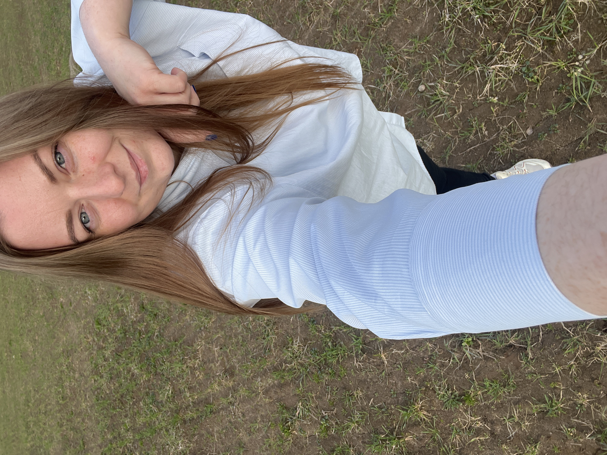 A young woman takes a selfie outdoors, with the ground visible below. The ground is patchy with grass and soil, suggesting an open field or park area. The woman has light brown hair and is wearing a light blue striped button-down shirt. She is smiling slightly and looking at the camera. The picture appears to be taken at arm's length, giving a close-up view of the woman and a wider view of the surrounding environment. The location is Razdolye, Russia.