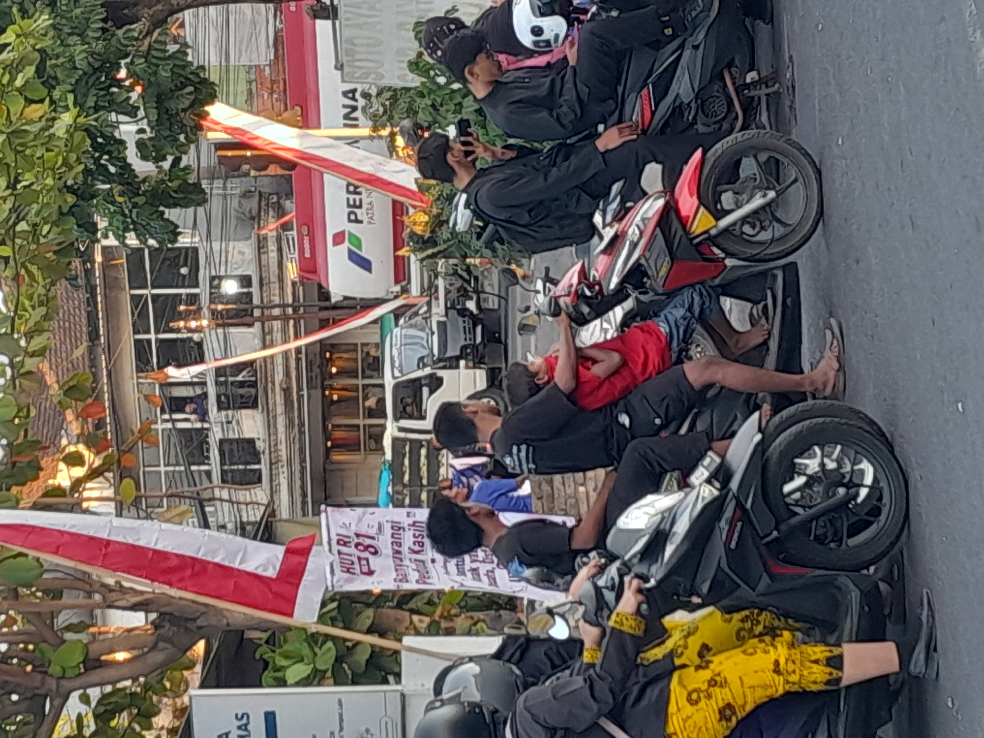 This is a medium-angle, eye-level, full shot of people on motorcycles in Gentengwetan, Indonesia. In the foreground, several individuals are seated on motorcycles. A person on the left is wearing a black helmet and yellow patterned pants. Next to them, a man in a black shirt and black pants sits on a motorcycle. Behind them, another man in a black shirt and black pants sits with a child in a red shirt. To the right, two men in black shirts and black pants are seated on motorcycles, with one holding a camera. In the background, a large white truck with red and white flags is visible, with the logo "PERTAMINA" on its side. A banner with "HUT RI 81" and Indonesian text hangs from a pole. The environment appears to be a street with buildings and trees. The lighting suggests it is daytime, possibly late afternoon.
