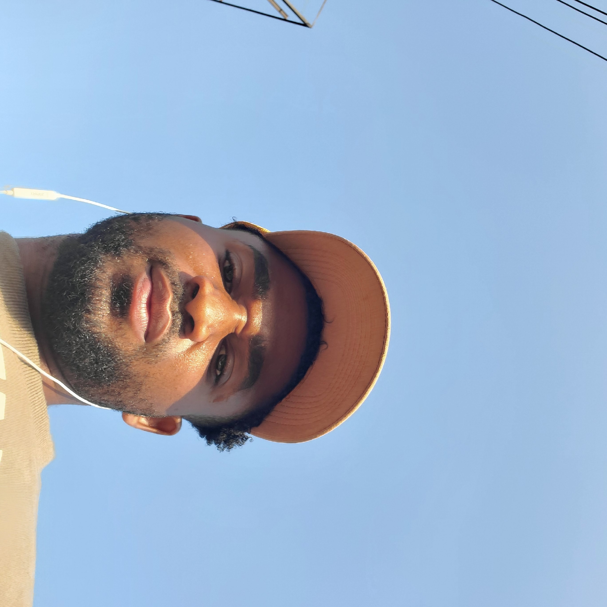 A person wearing a tan cap and earphones is looking up at the camera, with a clear blue sky in the background. There are utility lines in the upper portion of the image. The location in the city of Onitsha, Nigeria, cannot be determined from this image alone.