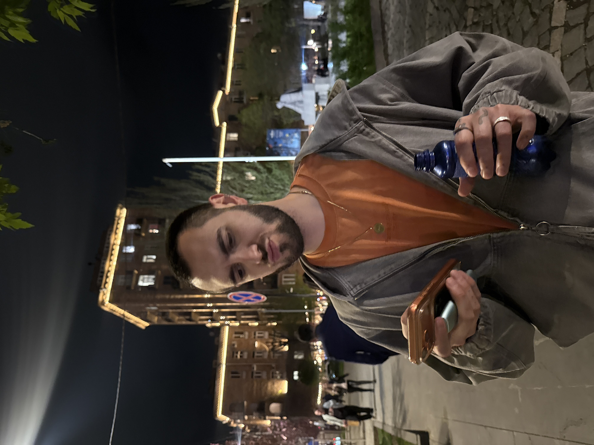 A person, presumably male, is holding a blue plastic water bottle in the left hand and a phone in the right hand. The individual has dark hair, stubble, and is wearing an orange shirt and a gray jacket.
The backdrop shows a street scene at night, potentially in Yerevan, Armenia. Architectural features with decorative lighting suggest an urban environment, with other people walking along the sidewalk. There is also a traffic sign visible.