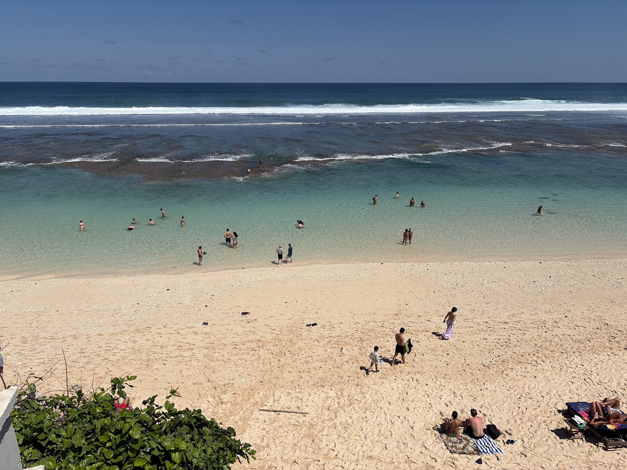 This aerial photograph captures a lively beach scene in Kuta Selatan, Indonesia, under a clear blue sky. The turquoise water of the ocean gently laps onto a wide expanse of golden sand. Numerous people are enjoying the beach and the water. Some are wading in the shallows, while others are swimming further out. A few individuals are walking along the shoreline, and others are relaxing on beach chairs and towels on the sand. In the foreground, a lush green bush with vibrant pink flowers grows near a white structure, suggesting a manicured area bordering the natural beach. The ocean displays distinct bands of color, with white waves breaking closer to shore, followed by clearer, lighter blue water over submerged reefs, and then a deeper blue further out. The beach is dotted with footprints, indicating recent activity, and several pairs of flip-flops are scattered on the sand.